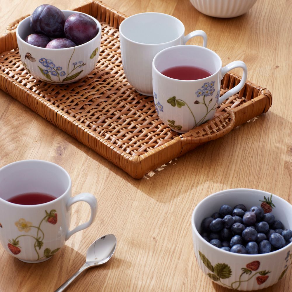 Ett träbord dukat med en rottingbricka med plommon, en enkel mugg och en mönstrad mugg med en röd dryck. På bordet finns också en annan mönstrad mugg med en röd dryck och en skål med blåbär.