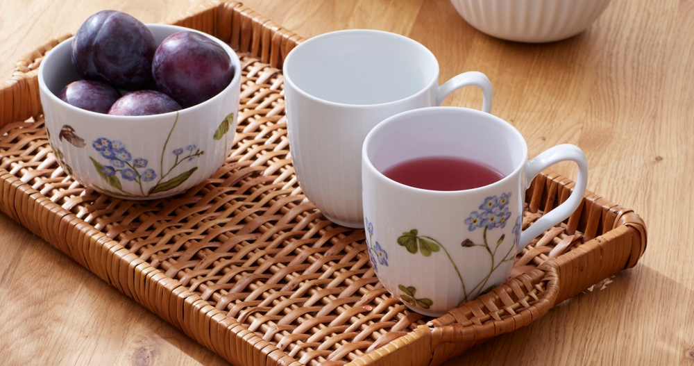 Ett träbord dukat med en rottingbricka med plommon, en enkel mugg och en mönstrad mugg med en röd dryck. På bordet finns också en annan mönstrad mugg med en röd dryck och en skål med blåbär.