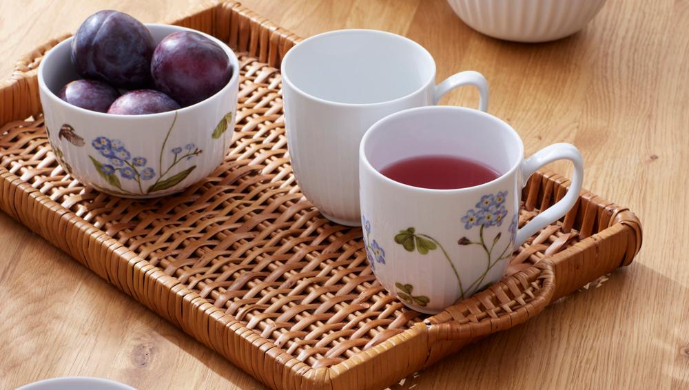 Dukat träbord med flätad bricka, blommiga porslinsmuggar med saft och skålar med plommon och blåbär.
