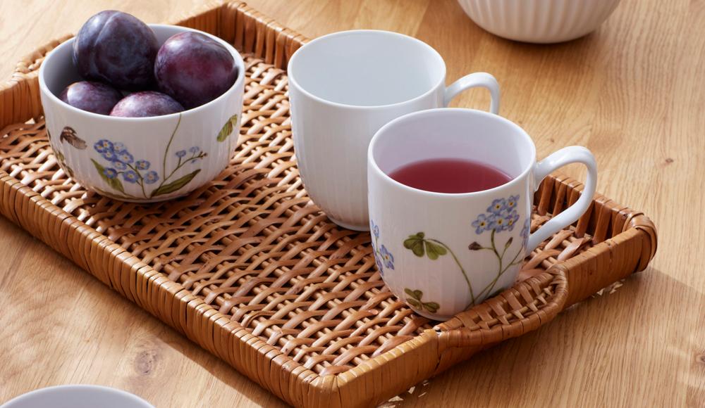 Ett träbord dukat med en rottingbricka med plommon, en enkel mugg och en mönstrad mugg med en röd dryck. På bordet finns också en annan mönstrad mugg med en röd dryck och en skål med blåbär.