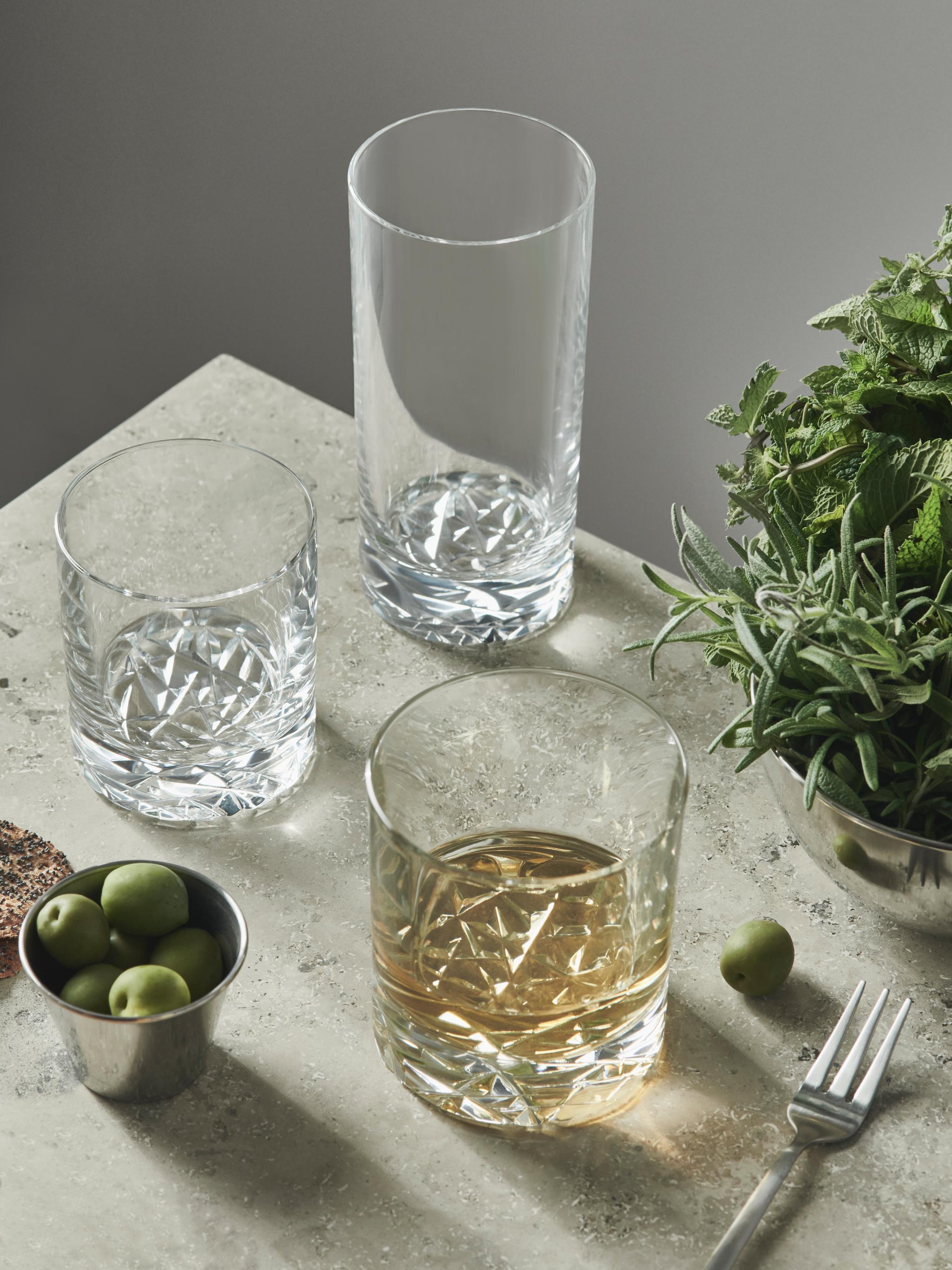 Tre dekorativa glas, ett med bärnstensfärgad vätska, står med gröna oliver, färska örter och en gaffel på en texturerad ljus yta.