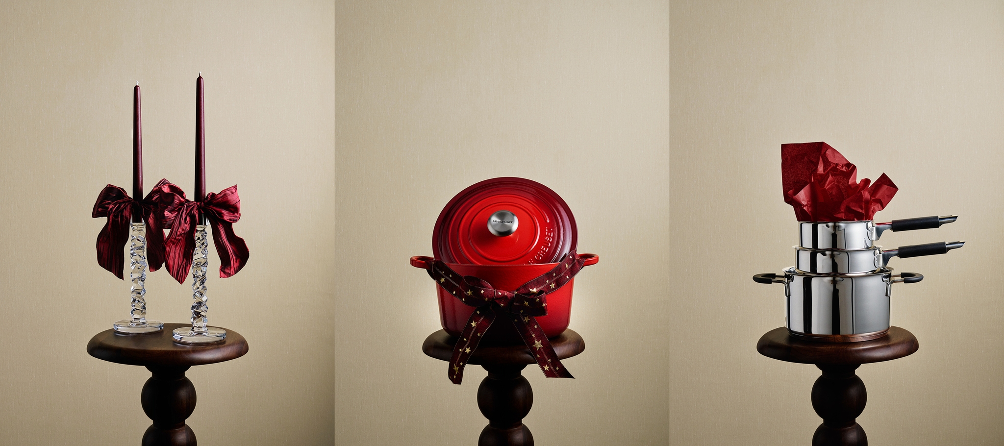 Kolme juhlavaa lahjaesinettä jalustoilla: punaiset kynttilät ruseteilla, punainen Le Creuset -ruukku rusetilla ja pinotut hopeiset ruukut punaisella nenäliinalla.