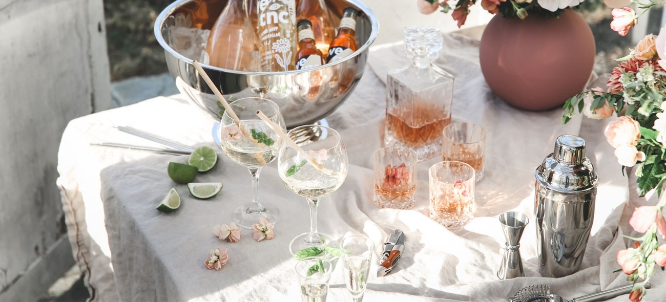 Shaker i silver på bord med vit duk och drinkar samt rosa blommor och lime