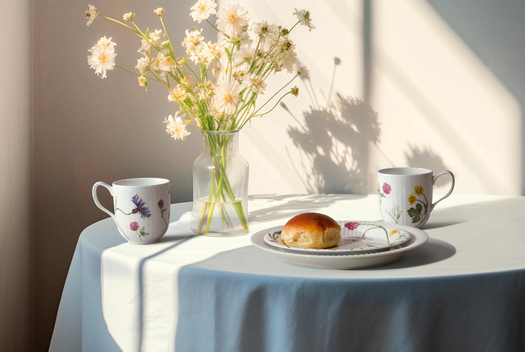 Ett bord med en vas med ljusa blommor, två blommiga muggar och en fralla, badande i solljus och skugga.