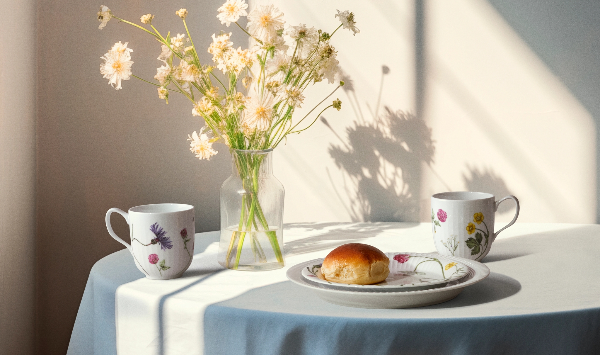 Ett bord med en vas med ljusa blommor, två blommiga muggar och en fralla, badande i solljus och skugga.