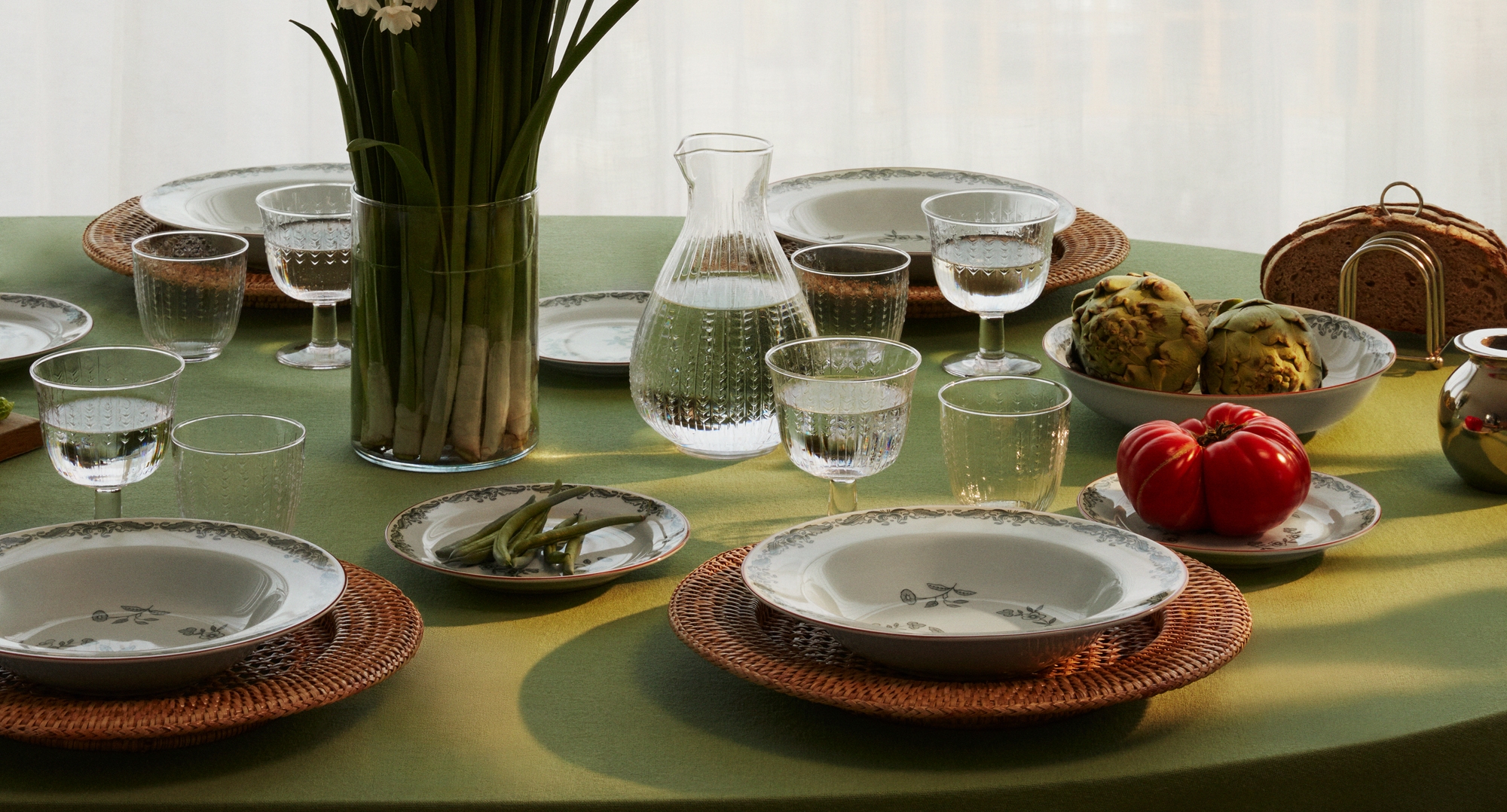 Vackert dukat matbord med grön duk, vita tallrikar, glas, en vas med vita blommor och färska grönsaker i mjukt dagsljus.