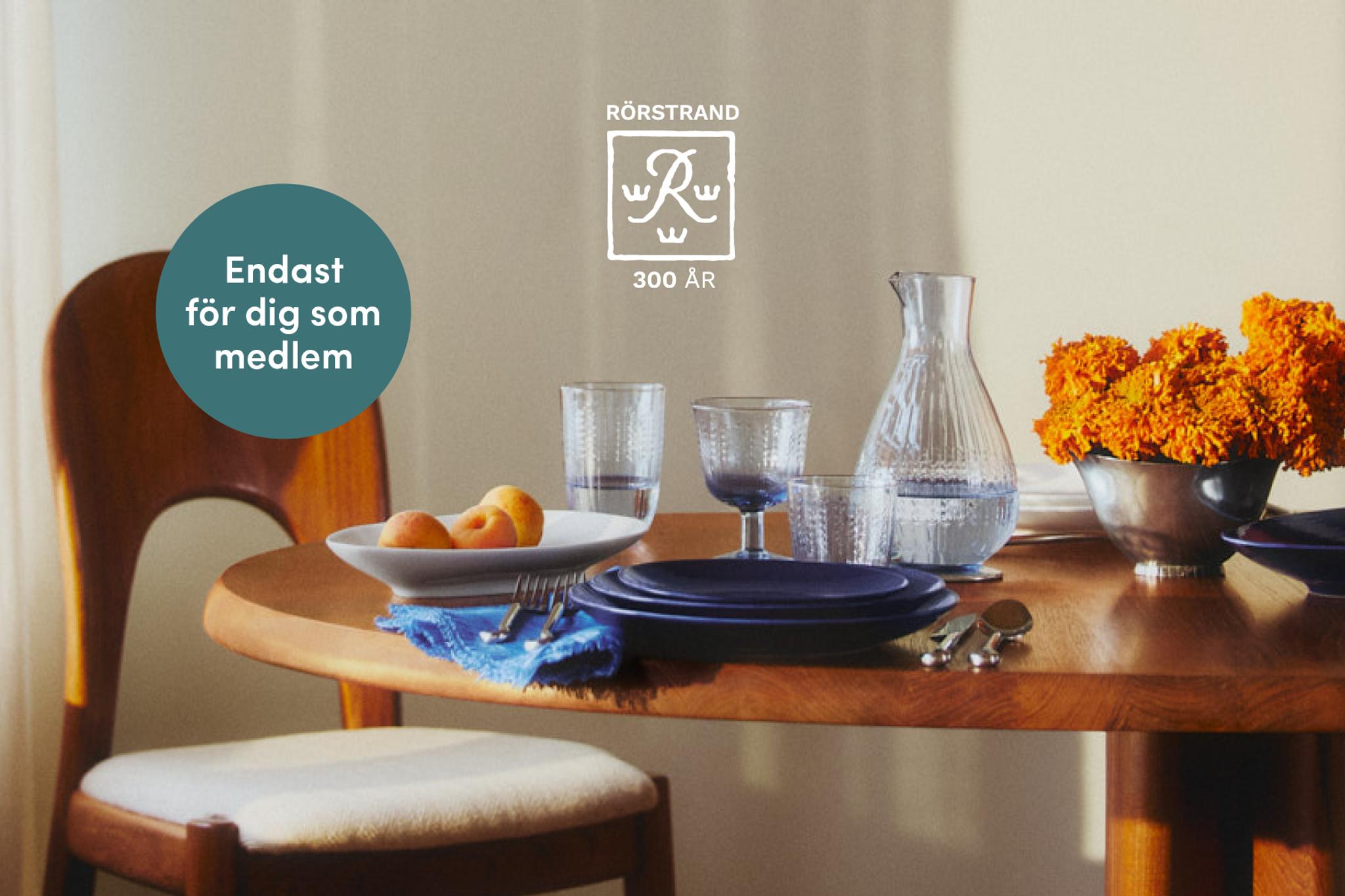 Ett matbord med mörkblå tallrikar, texturerade glas, en vattenkaraff, orange blommor och aprikoser. Text: RÖRSTRAND 300 ÅR och "Endast för dig som medlem".