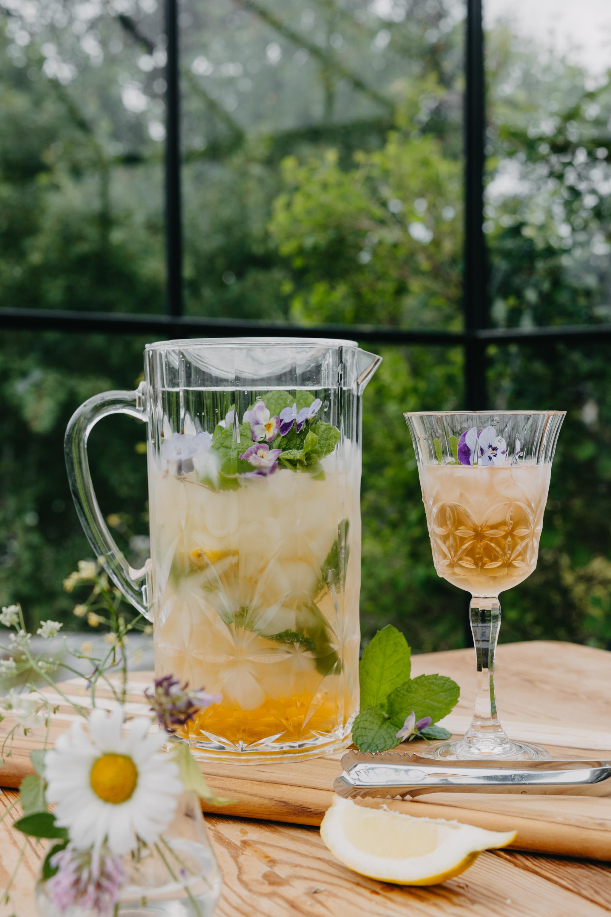Karaff och glas med somrig isdrink med citron, mynta och ätbara blommor på ett träbord i trädgården.