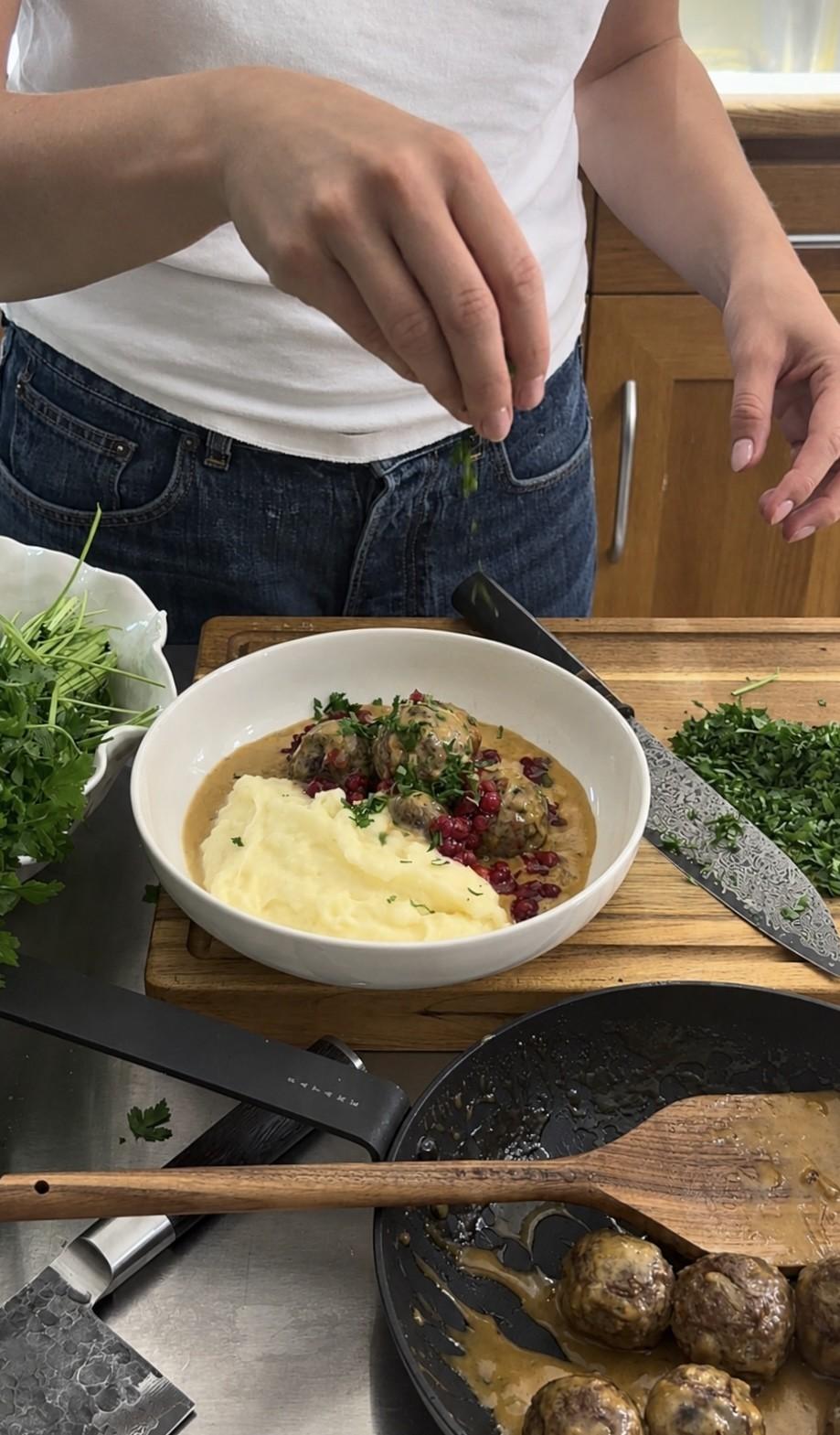 En person garnerar en skål med köttbullar, potatismos, gräddsås och lingon med färska örter.