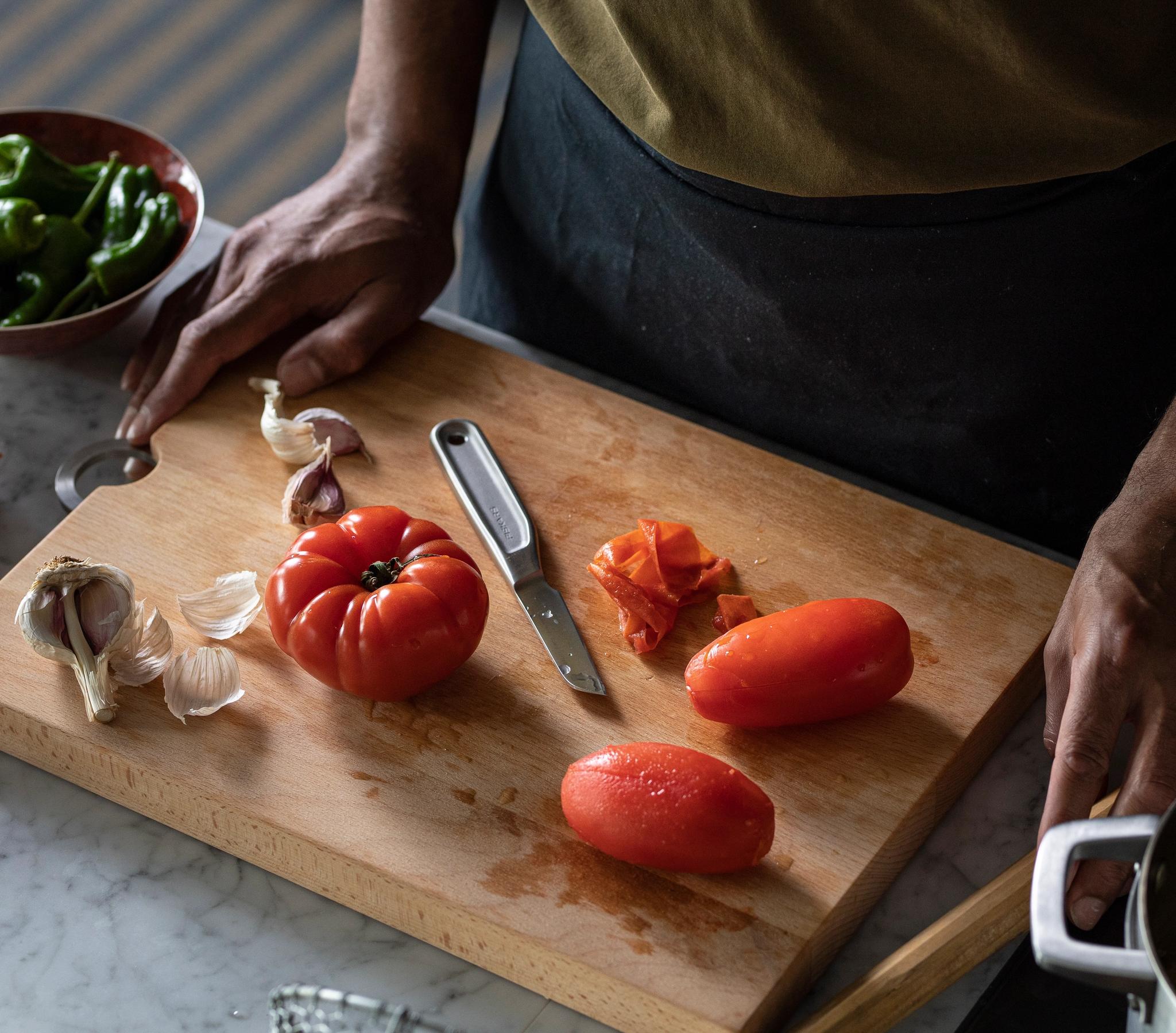 Kädet valmistavat tomaatteja ja valkosipulia puisella leikkuulaudalla, vihreiden paprikoiden, leivän ja ruukun lähellä.