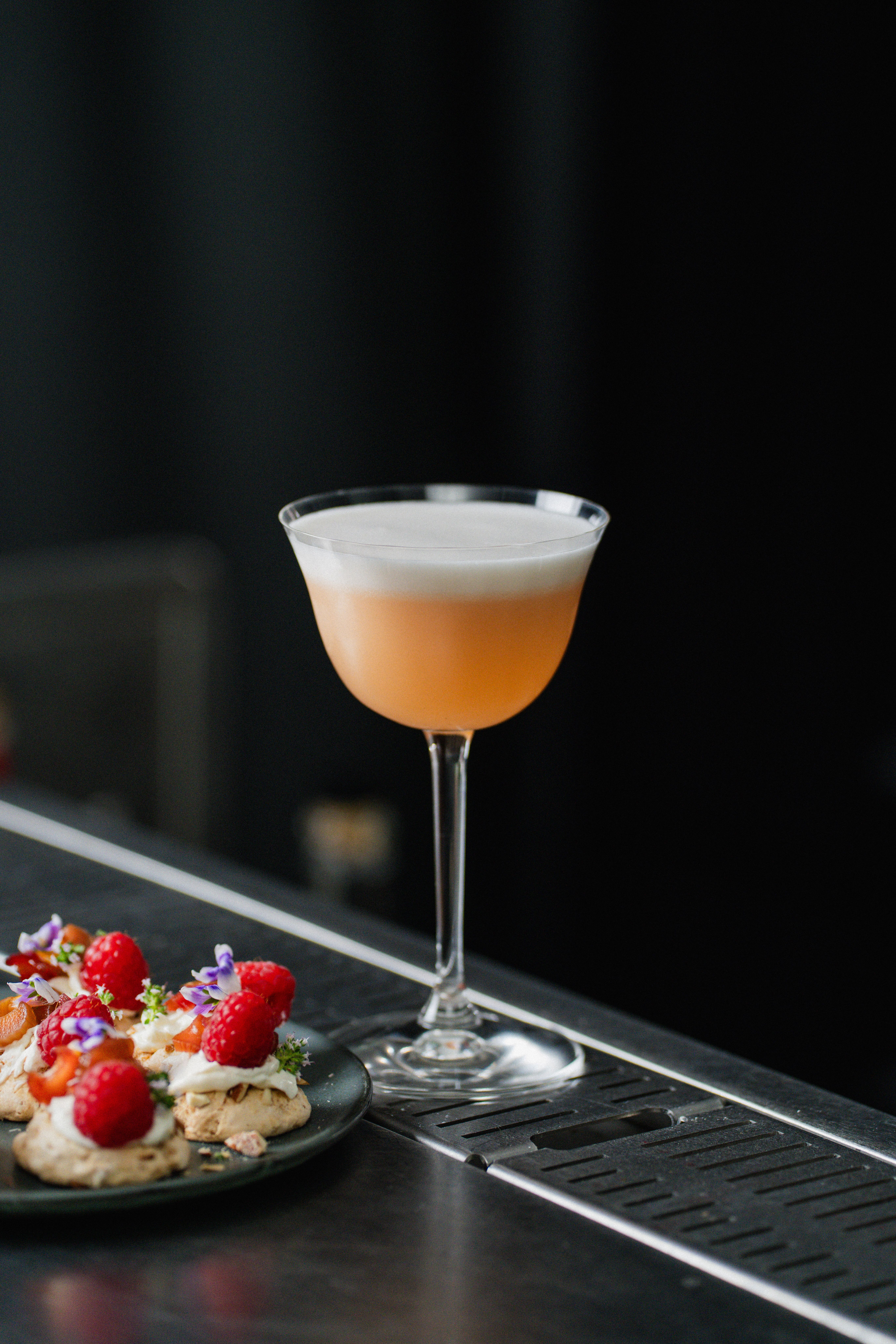 Elegant cocktail med skum serverad på bardisk bredvid små desserter med hallon och ätbara blommor.