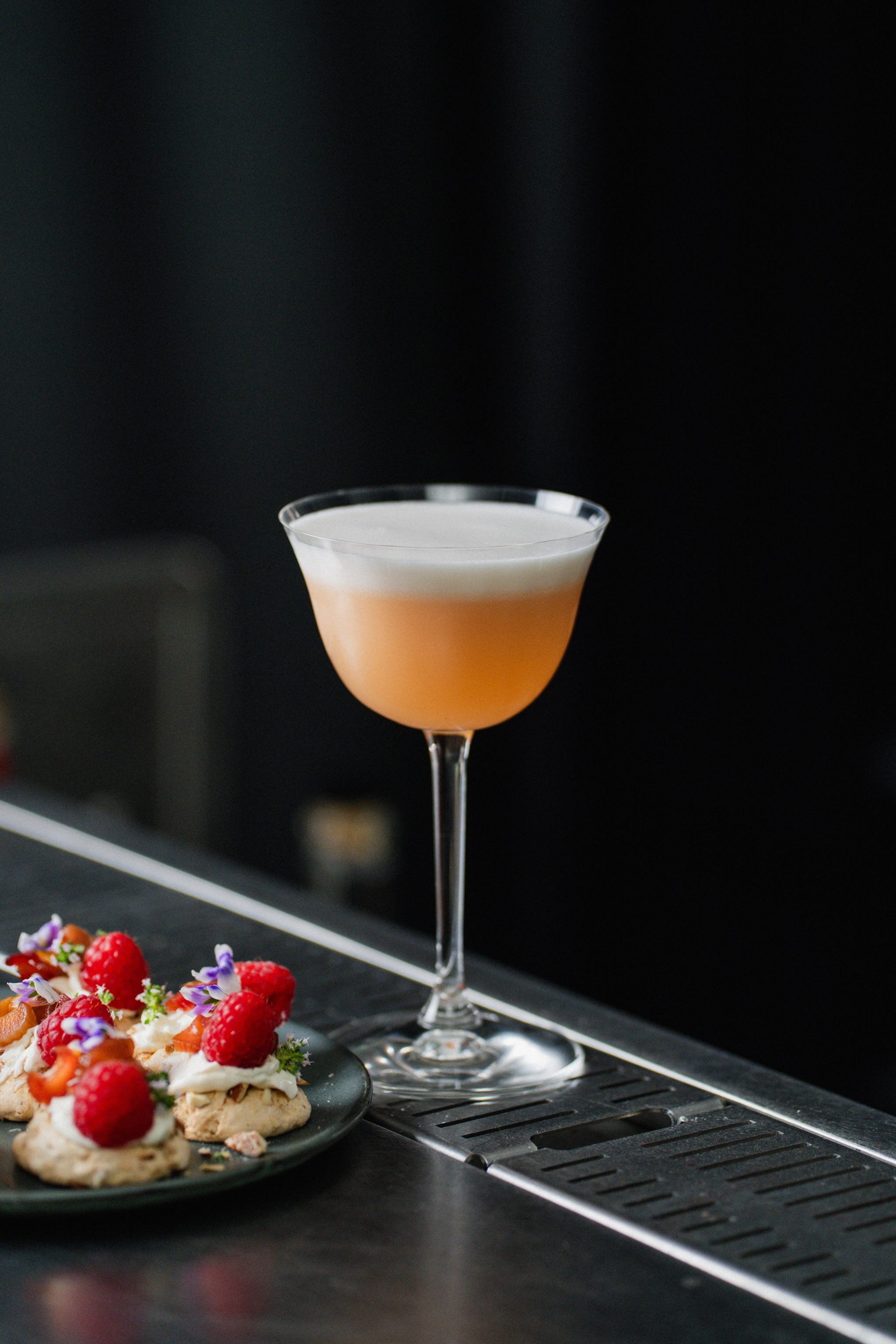 Elegant cocktail med skum topp och små desserter med hallon och ätbara blommor på ett svart fat i barmiljö.
