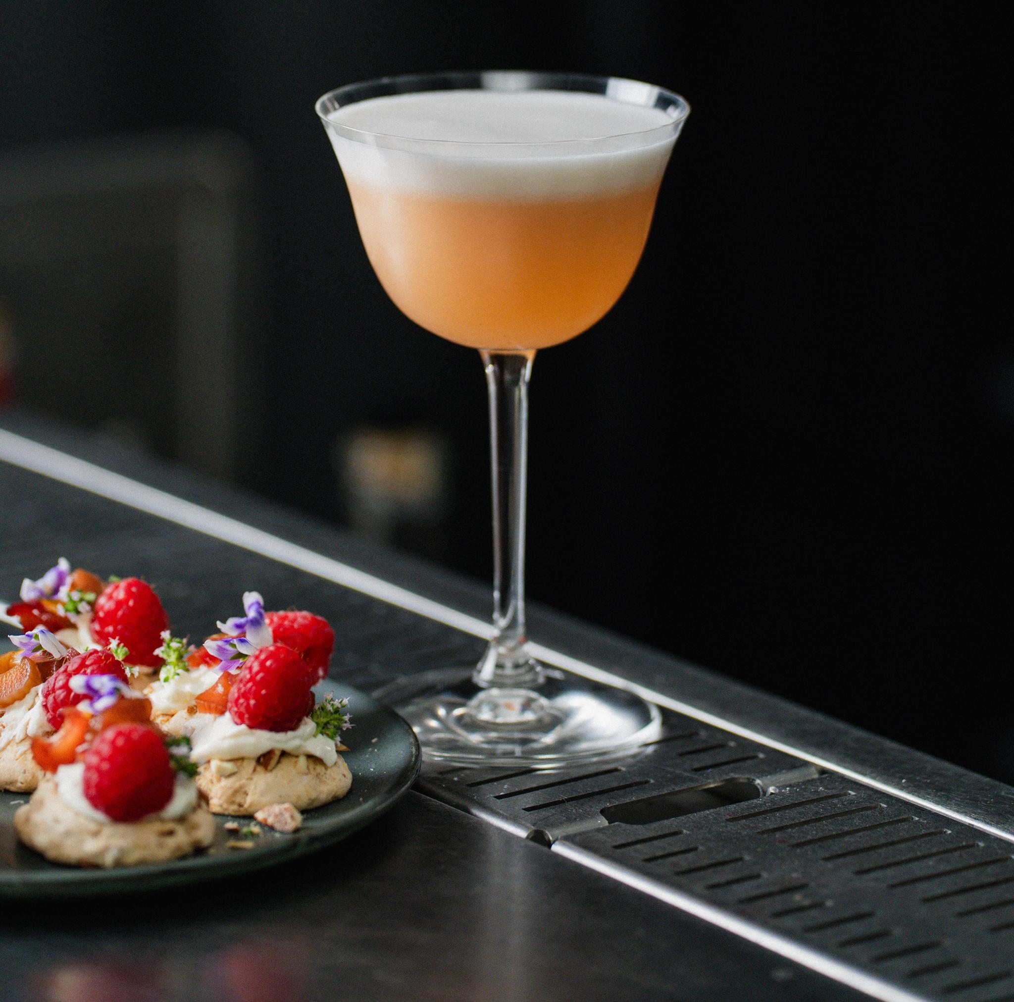 Elegant cocktail med skum serverad på bardisk bredvid små desserter med hallon och ätbara blommor.
