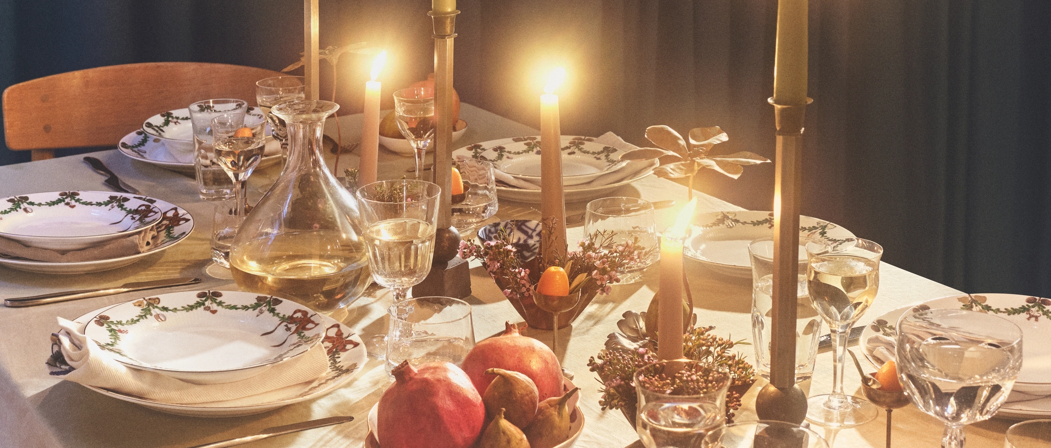 A festive dining table with glowing candles, decorative plates, a decanter, and pomegranates.