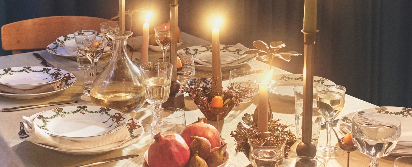 Lämpimästi valaistu ruokapöytä, jossa on juhlavia lautasia, hehkuvia kynttilöitä ja granaattiomenoita.
