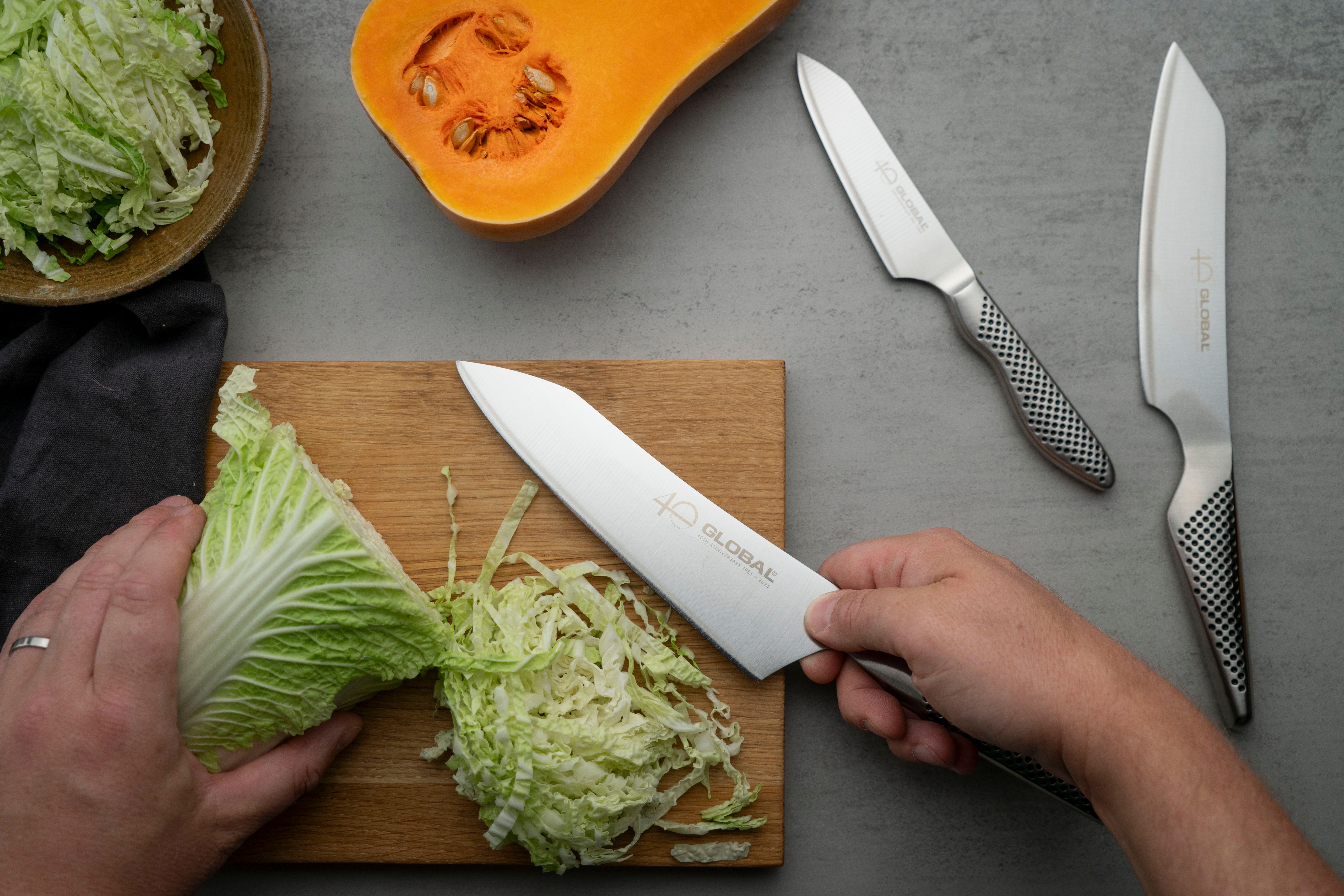 Hands chopping napa cabbage with a Global knife on a cutting board, with a halved butternut squash and other Global knives nearby.