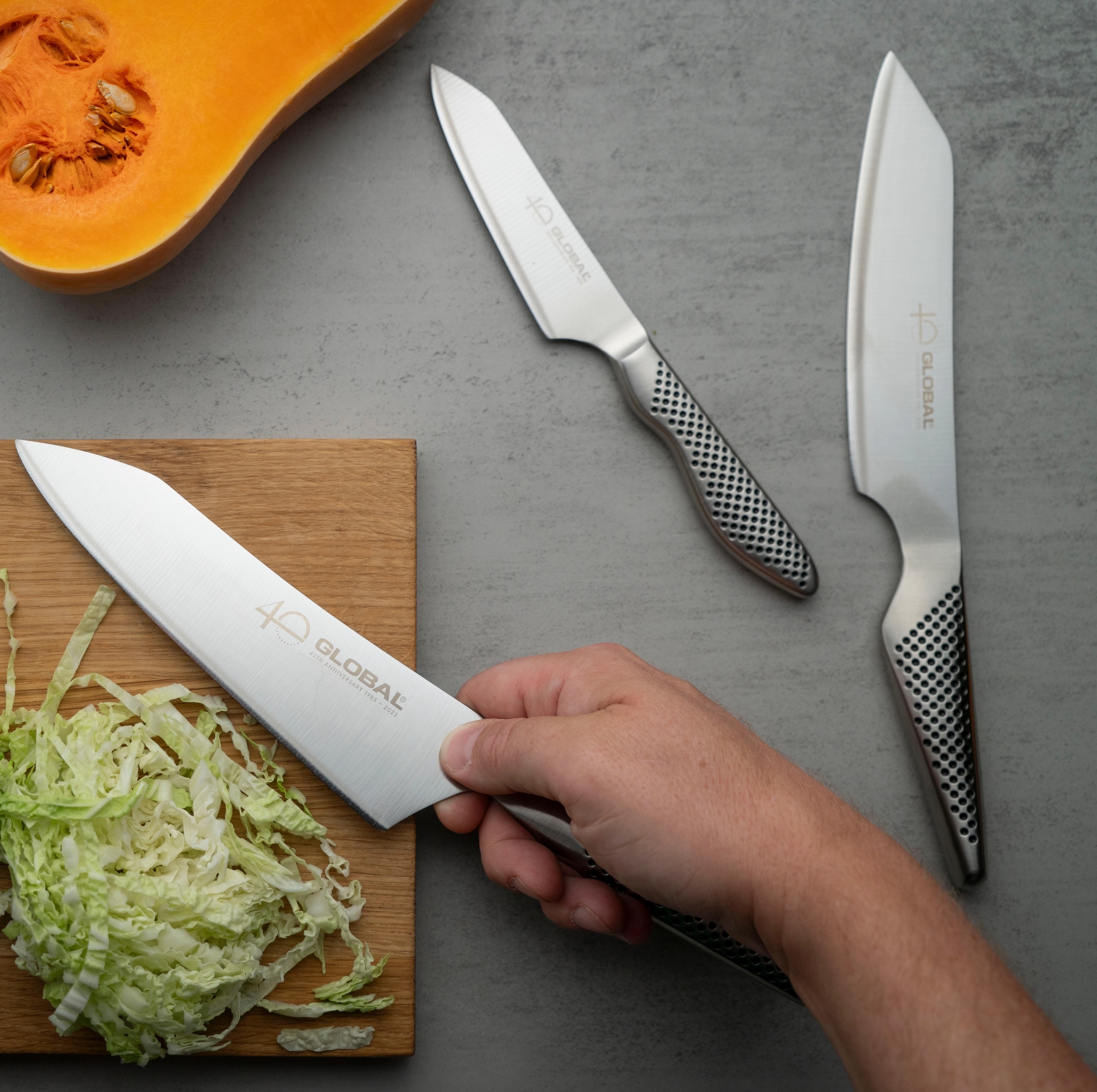 Overhead view of hands chopping Napa cabbage with a Global knife on a wooden board, with more knives and a butternut squash on a gray counter.