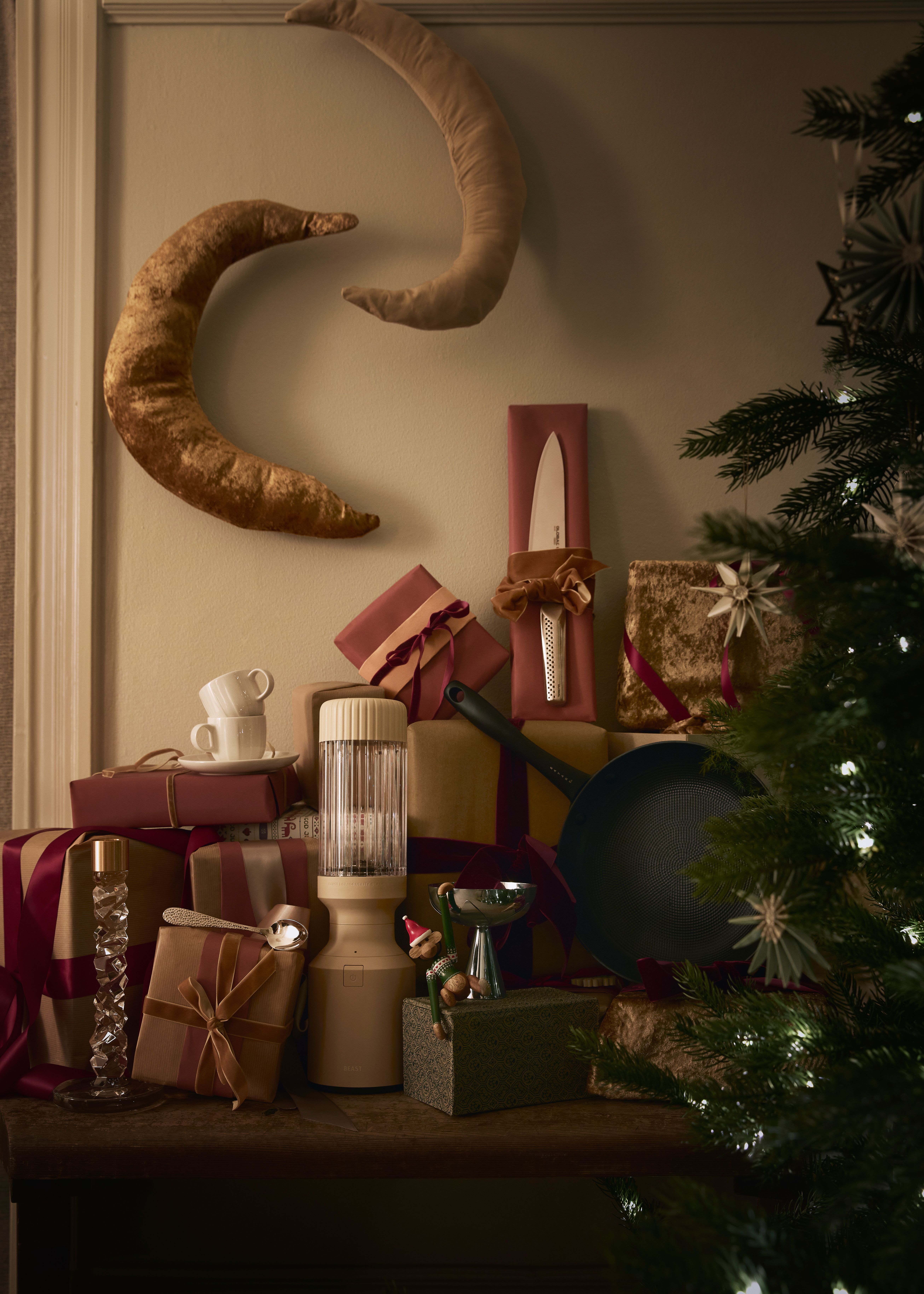 En festlig juleutstilling med innpakkede gaver og kjøkkenutstyr, inkludert en kniv, panne og blender, arrangert på en overflate med dekorative halvmåner og et juletre.