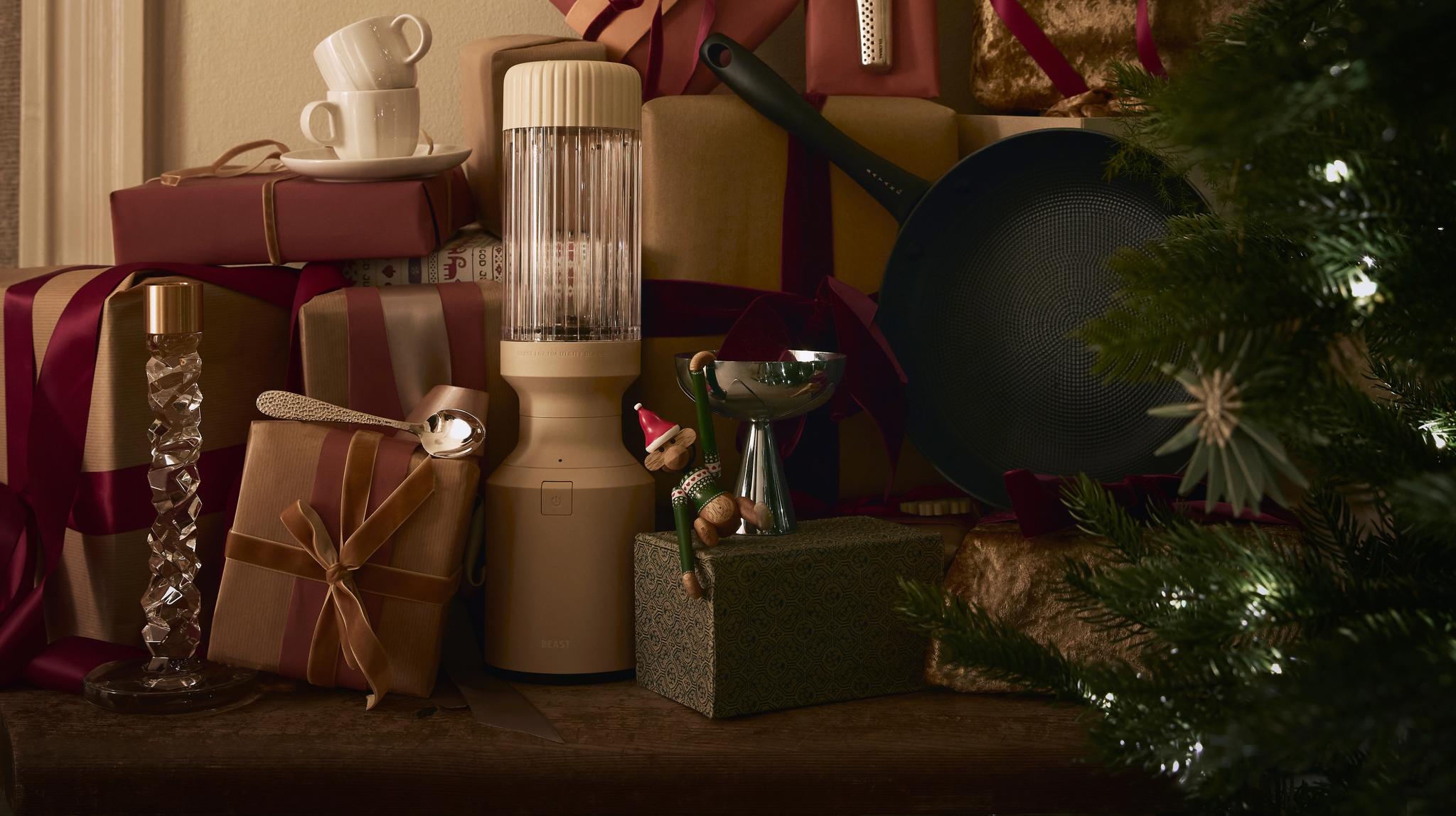 Kodikas asetelma, jossa on runsaasti joululahjoiksi paketoituja paketteja – mukana myös tehosekoitin ja paistinpannu – pöydällä kahden kuunsirppikoristeen alla. Oikealla näkyy osittain koristeltu joulukuusi.