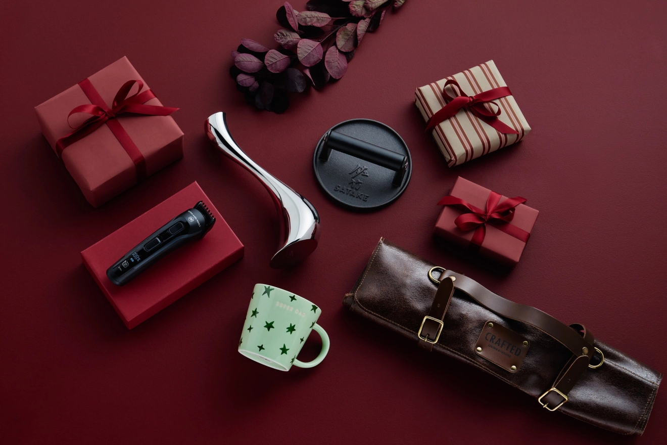 A flat lay of various items on a deep red background: wrapped gift boxes, a beard trimmer, a facial massage tool, a cast iron press, a green mug, and a leather case.