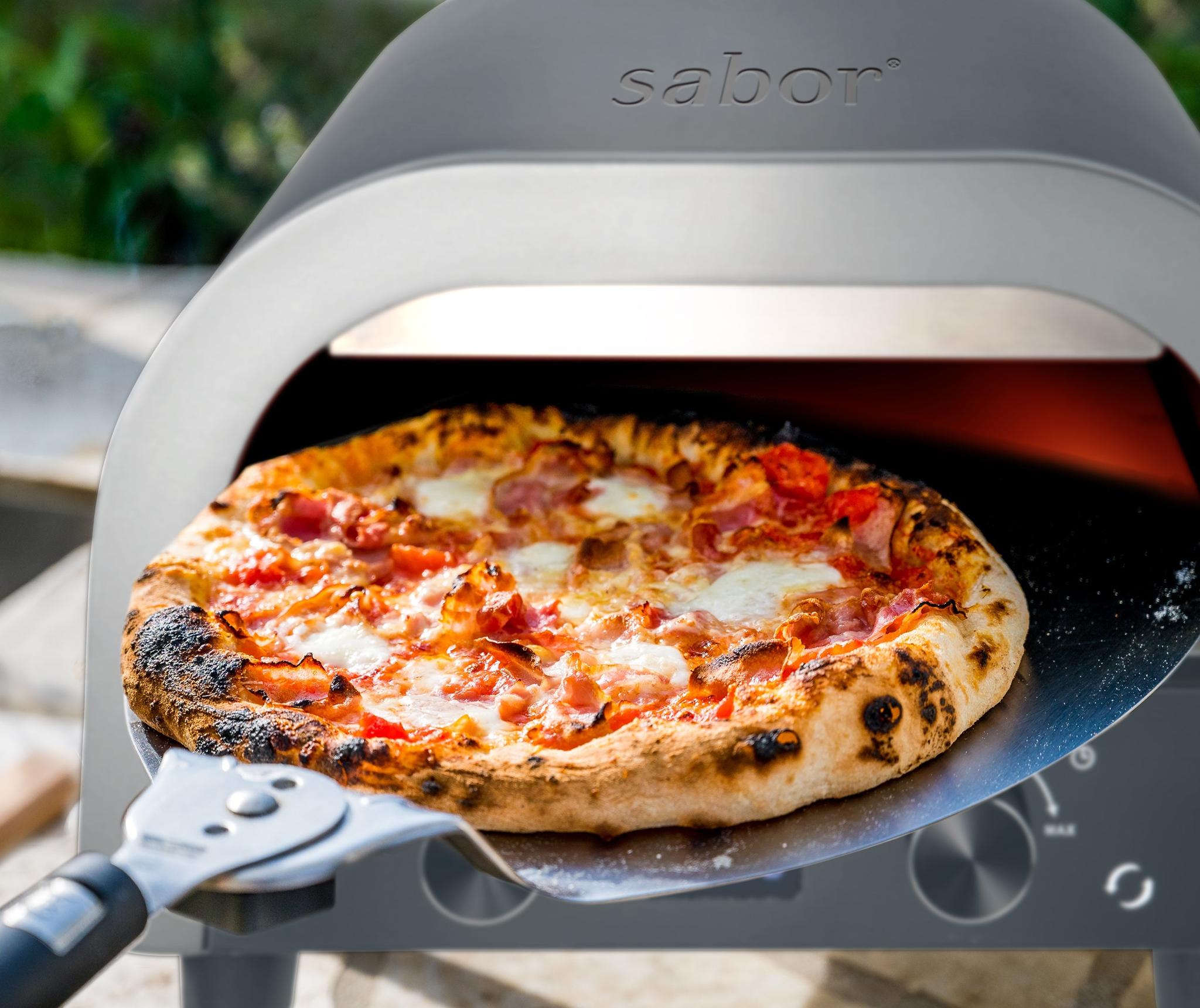 A freshly baked pizza with melted cheese and a bubbly crust being removed from a Sabor pizza oven with a metal peel.