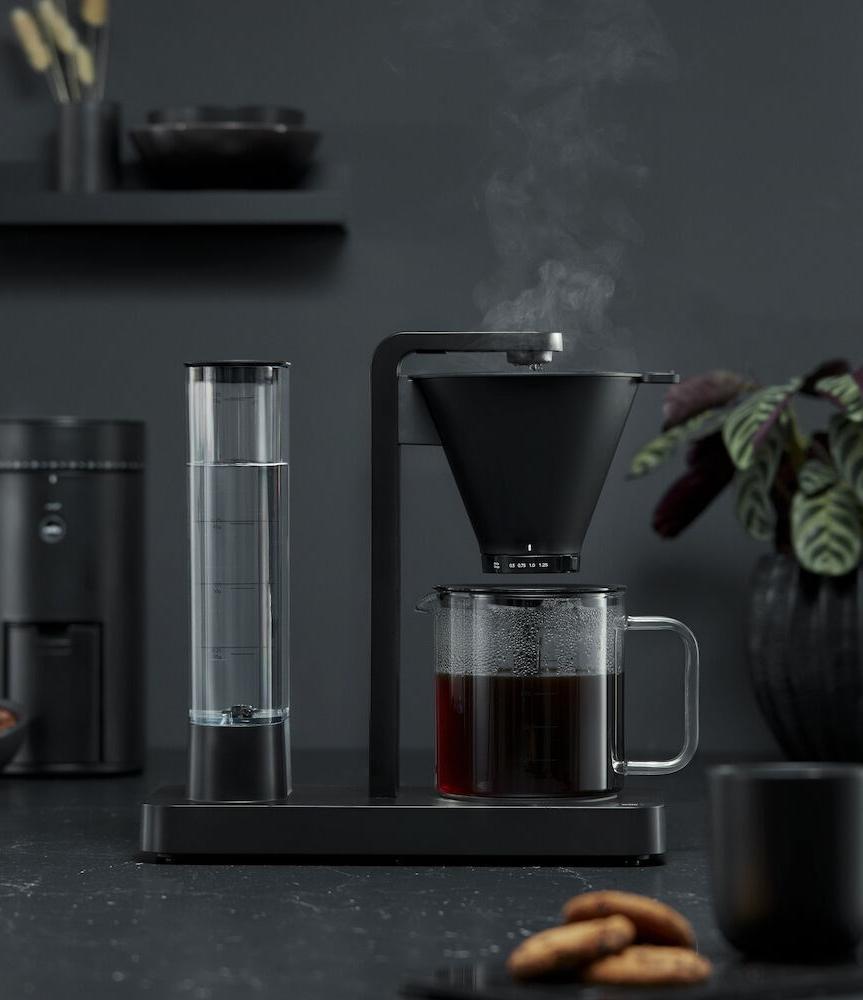 A minimalist black coffee maker brewing hot coffee into a glass carafe, with steam rising.