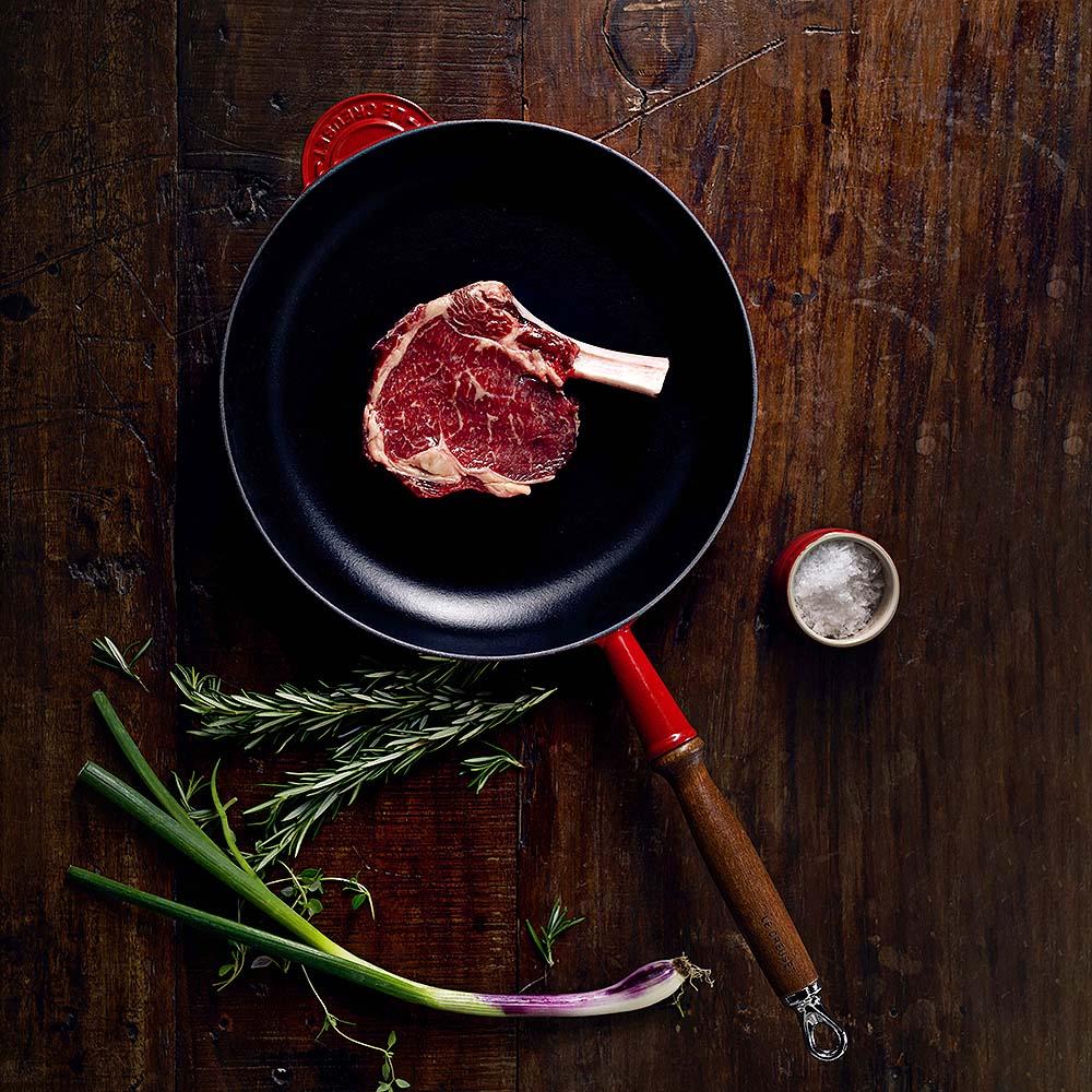 Rå ribeye-biff i en svart panna med örter och salt på en träbakgrund.