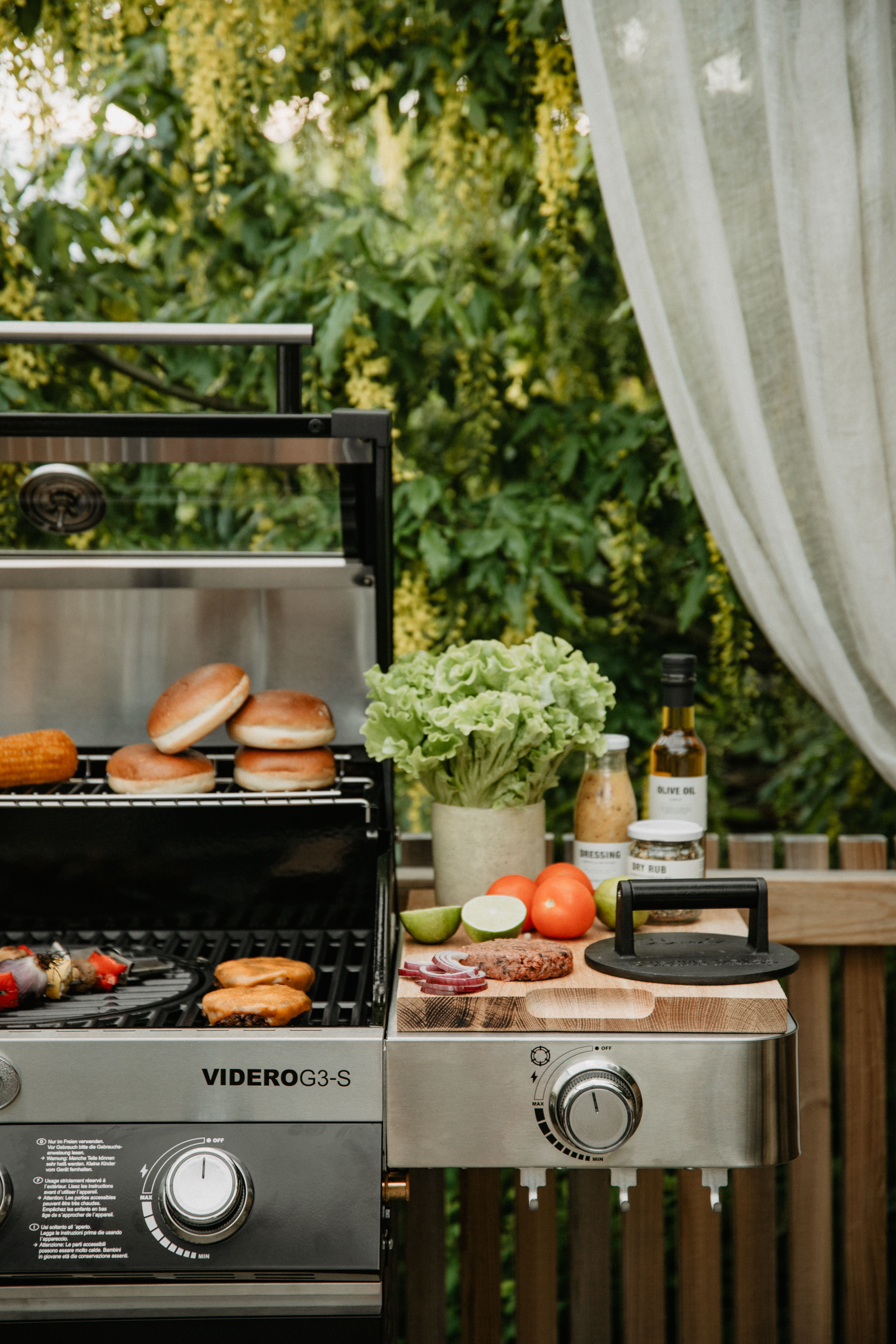 En utomhusgasgrill som lagar hamburgarbröd och majs, med färska hamburgaringredienser på en skärbräda av trä på sitt sidobord.