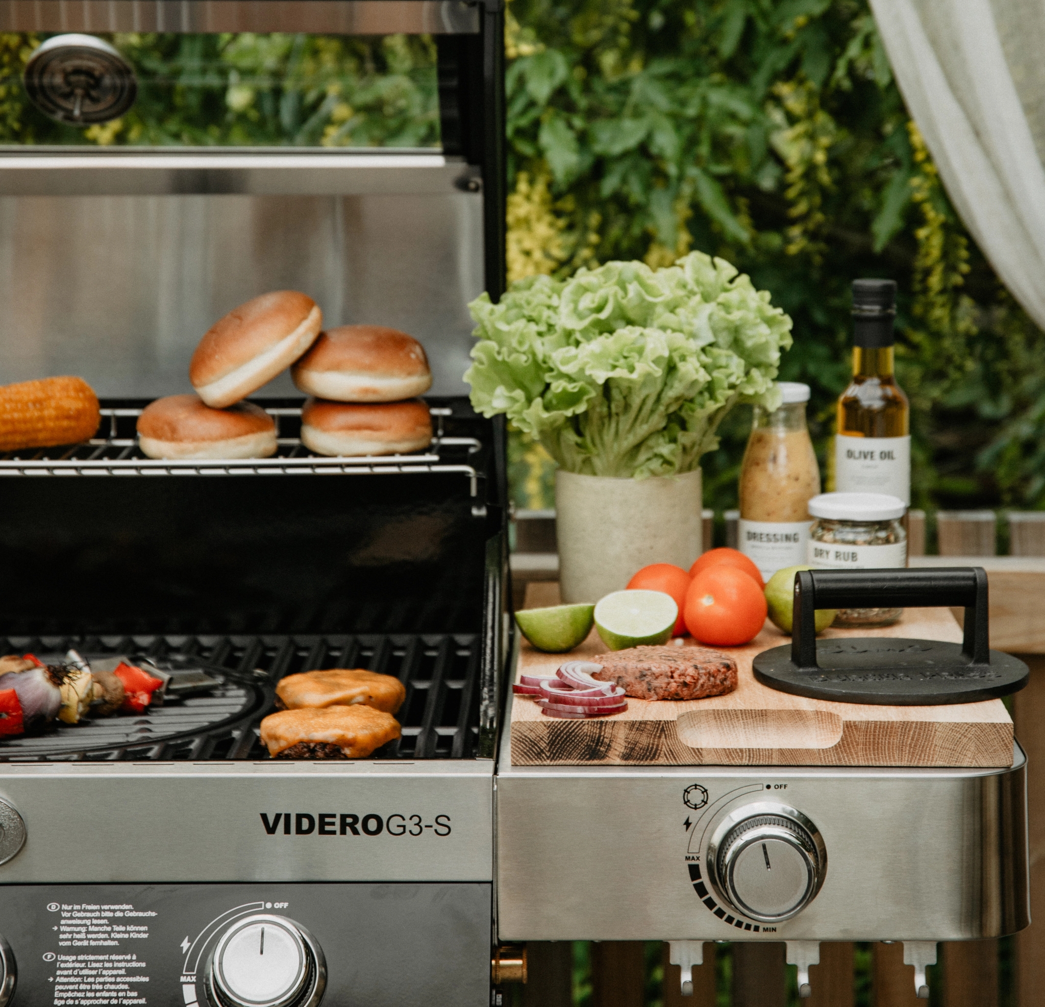Somrig utomhusgrill med hamburgare, bröd och grönsaker redo för grillning på en altan omgiven av grönska.