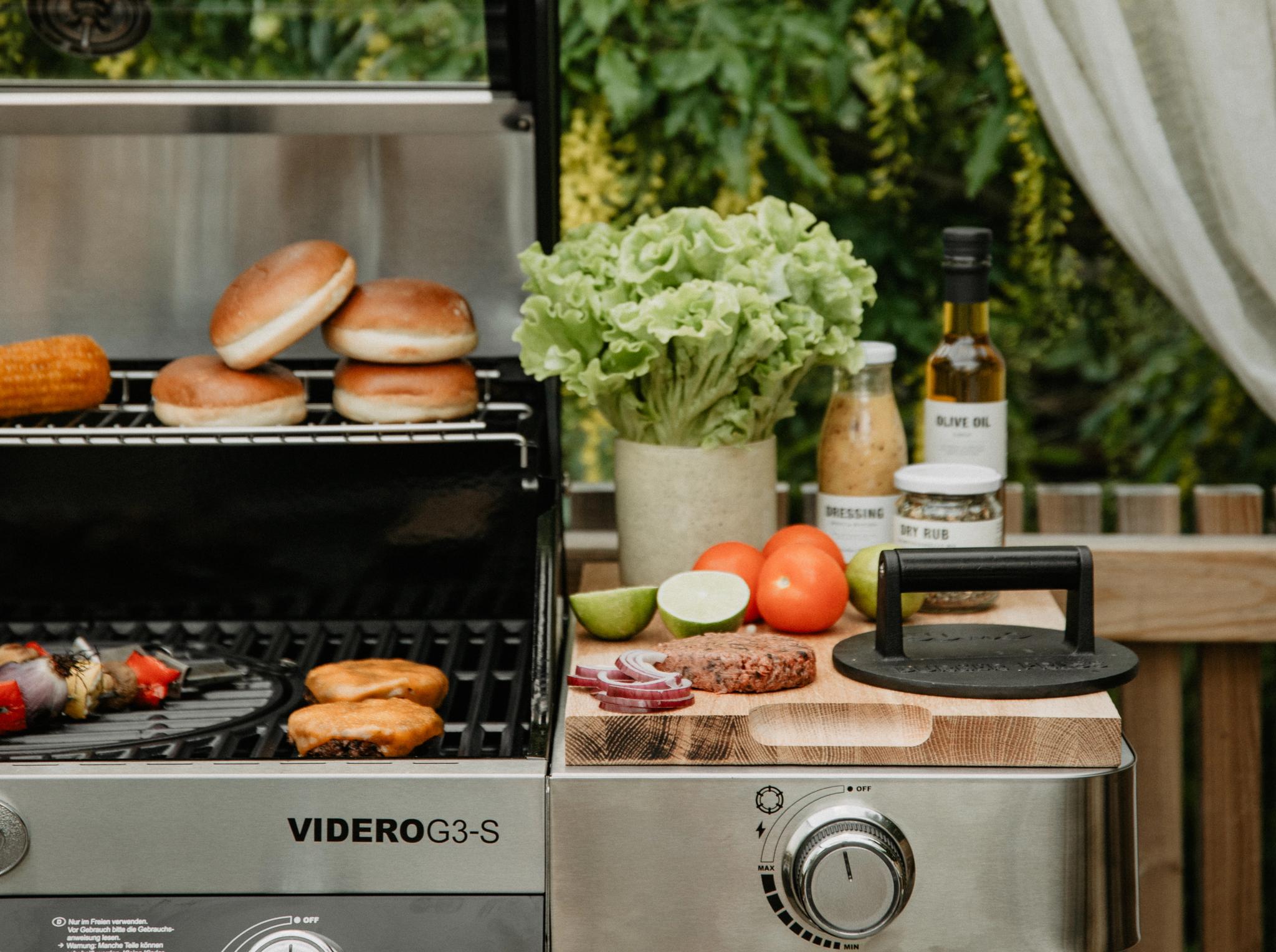 Somrig utomhusgrill med hamburgare, bröd och färska grönsaker uppdukade på en träbänk i grönskande trädgårdsmiljö.