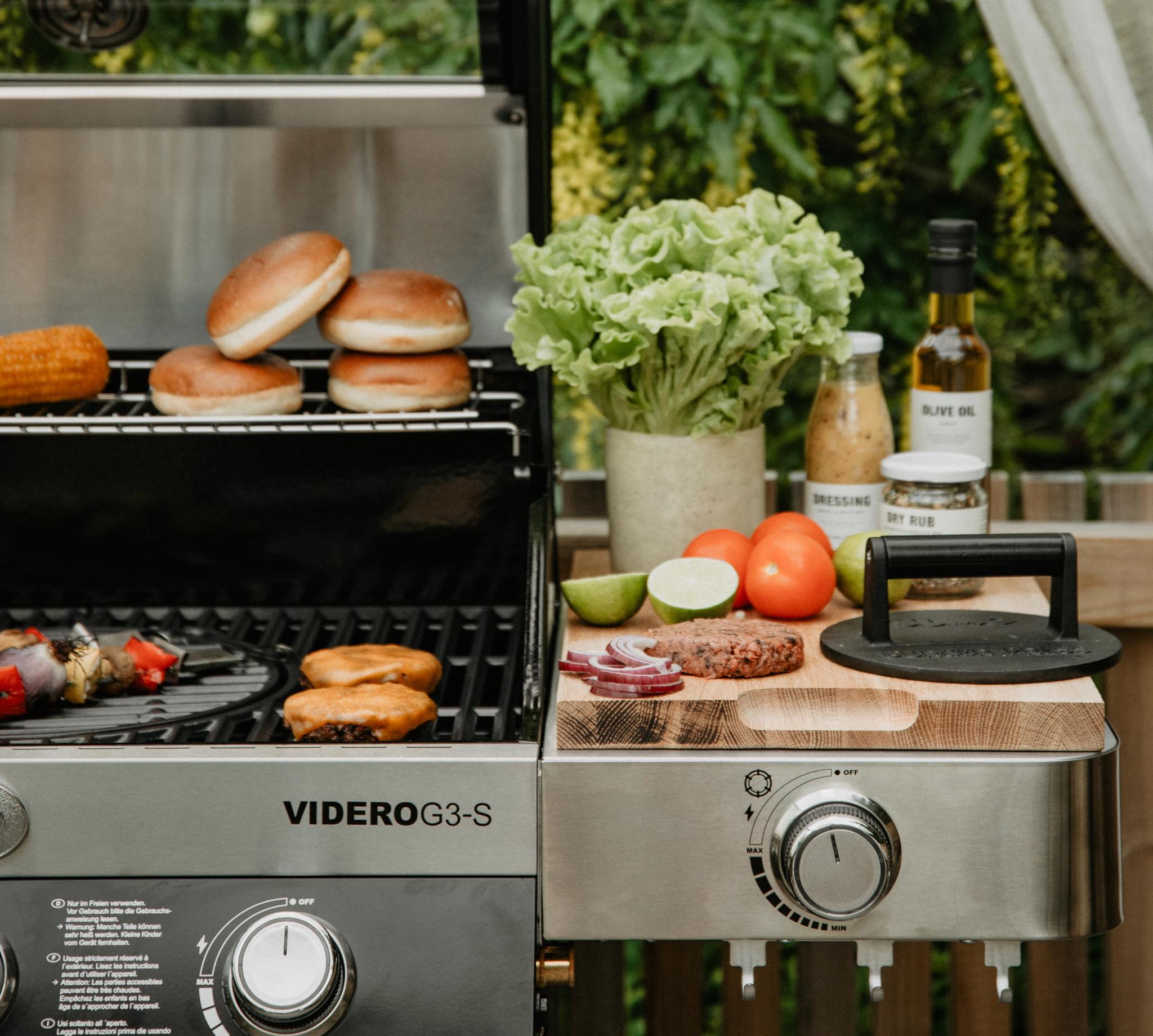 Somrig utomhusgrill med hamburgarbröd, grönsaker och tillbehör uppdukade på en altan framför grönskande träd.