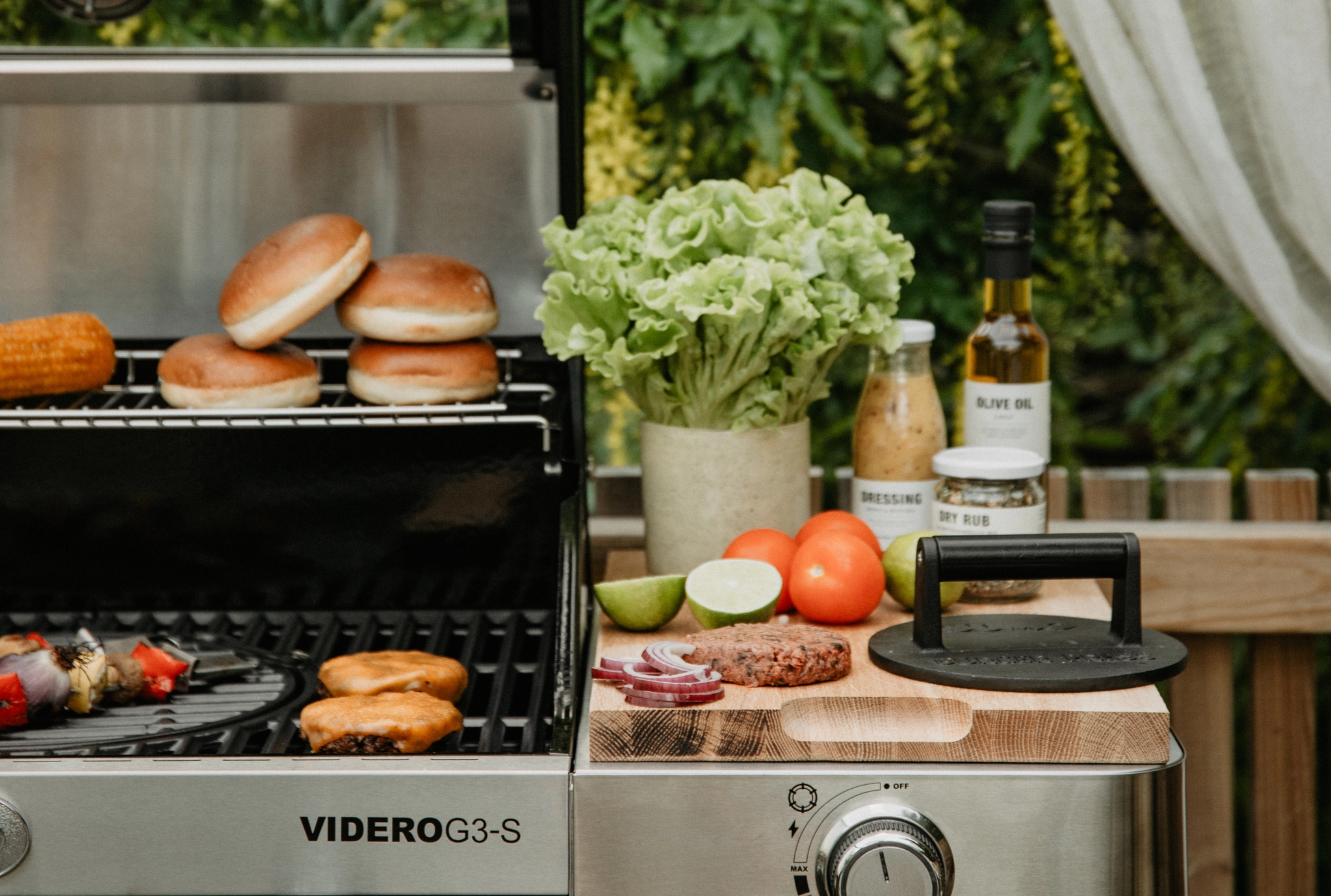 Somrig utomhusgrill med hamburgarbröd, grönsaker och tillbehör uppdukade på en modern gasolgrill på altanen.
