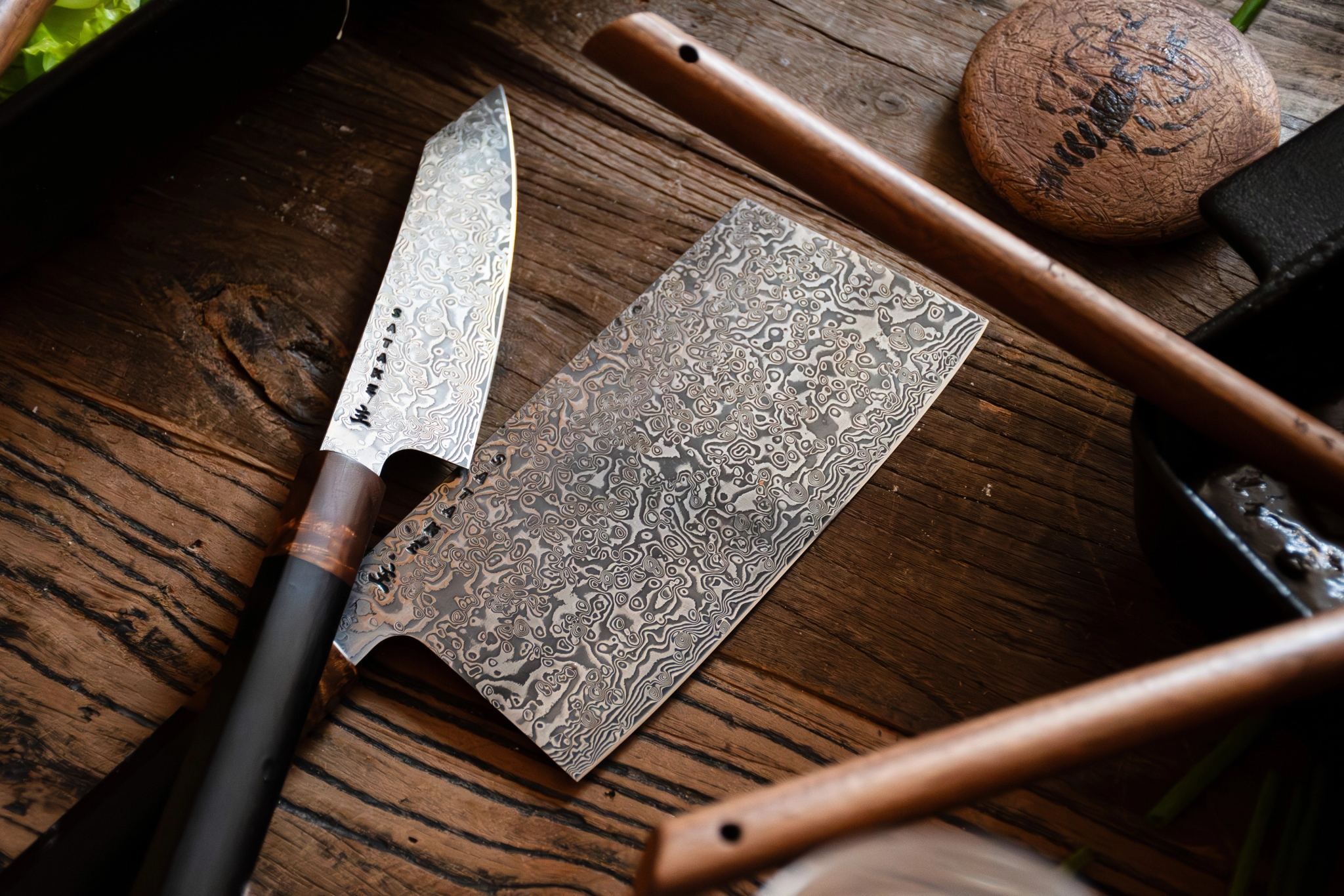 Två damaskusstålknivar och köksredskap på en rustik träyta.