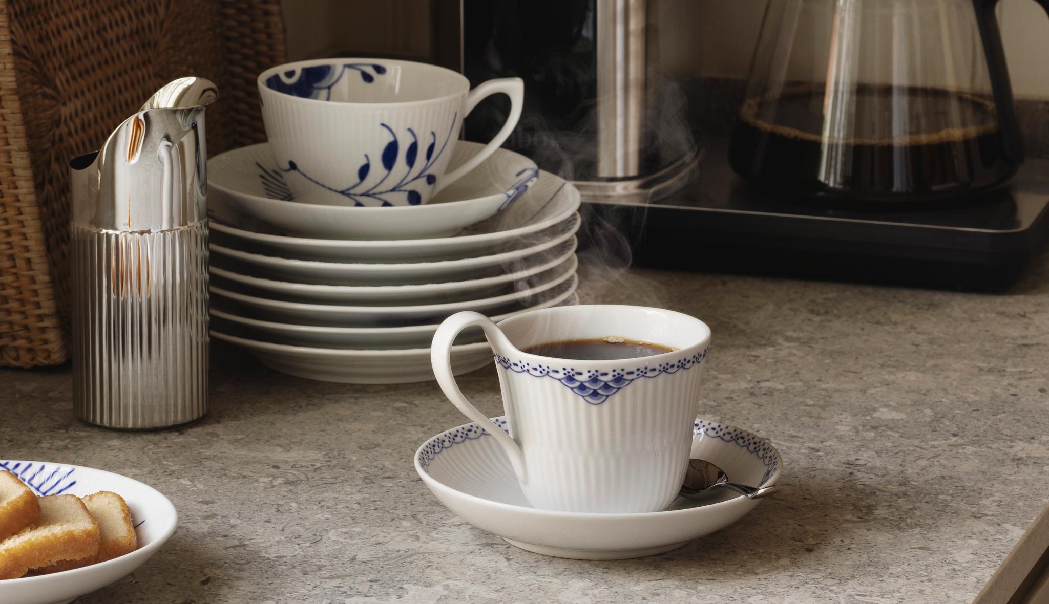 Höyryävää kahvia valkoisissa posliinikupeissa, joissa on sinisiä kukkakuvioita, ja pino yhteensopivia lautasia harmaalla tiskillä.