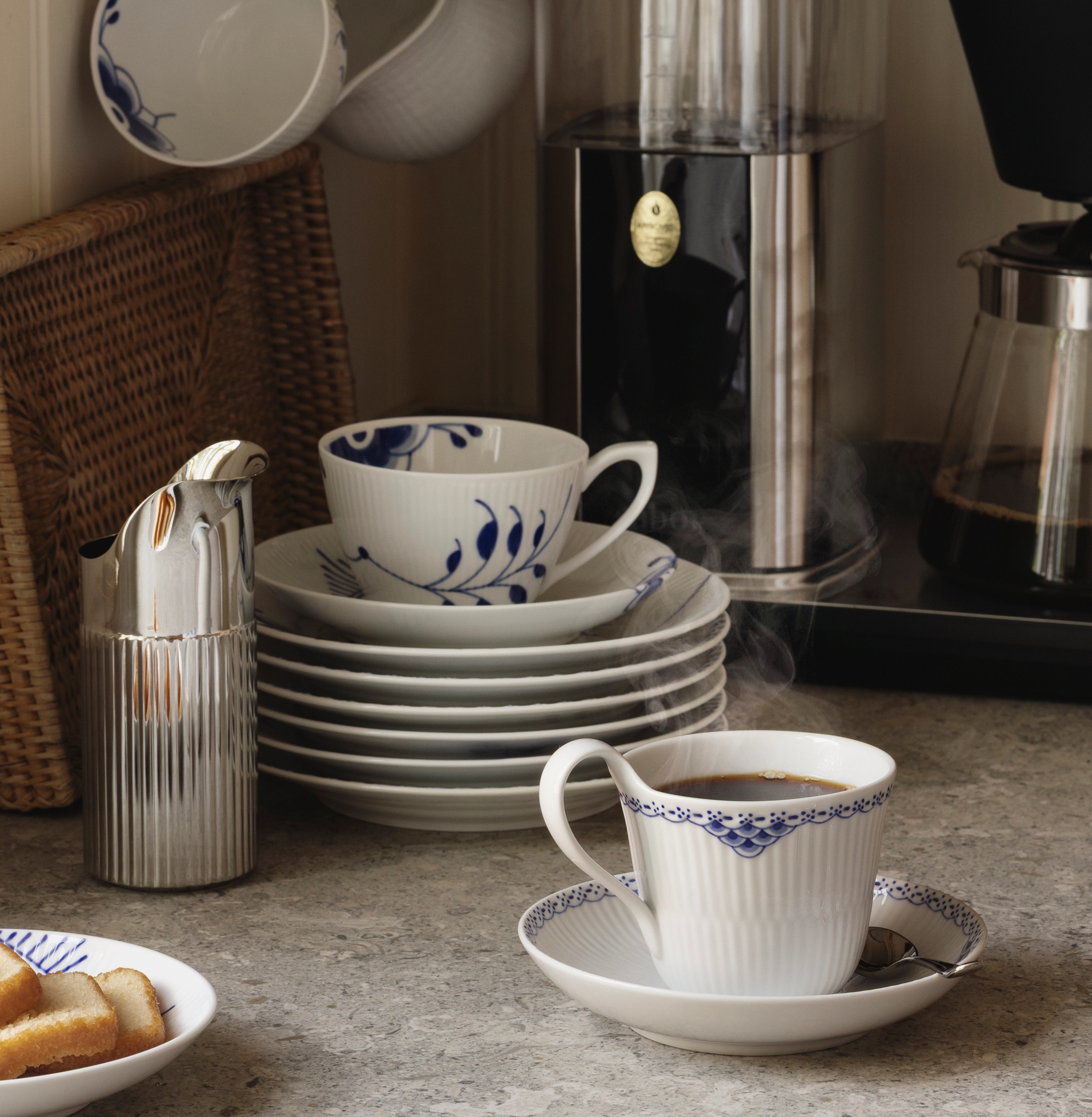 Höyryävää kahvia valkoisissa posliinikupeissa, joissa on sinisiä kukkakuvioita, ja pino yhteensopivia lautasia harmaalla tiskillä.
