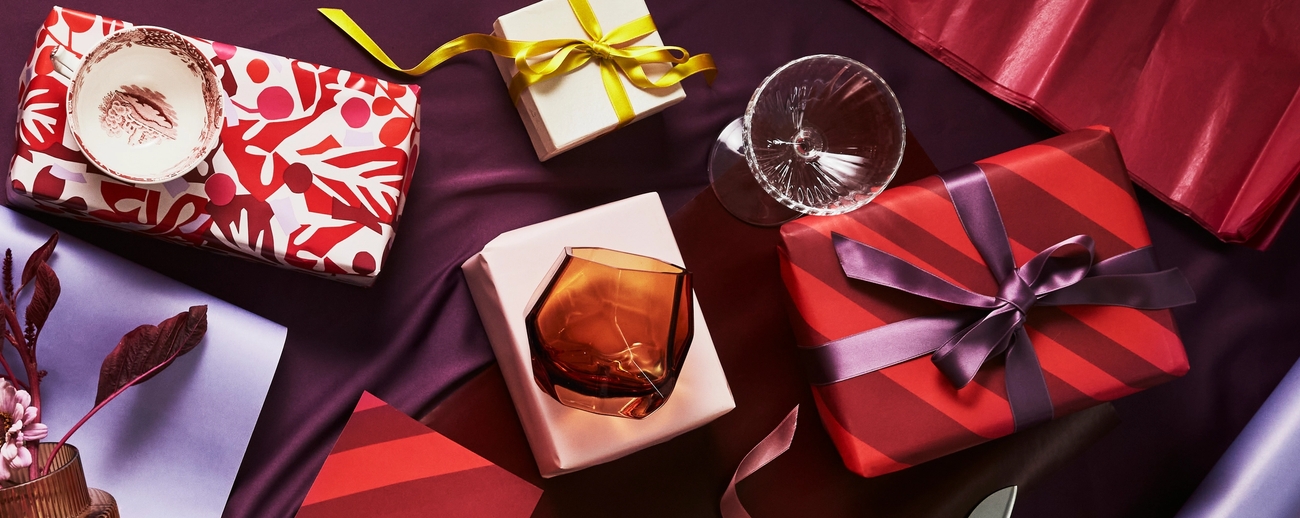 Gaveinnpakkede esker, glass og gavepapir i festlige røde, lilla og gulltoner.