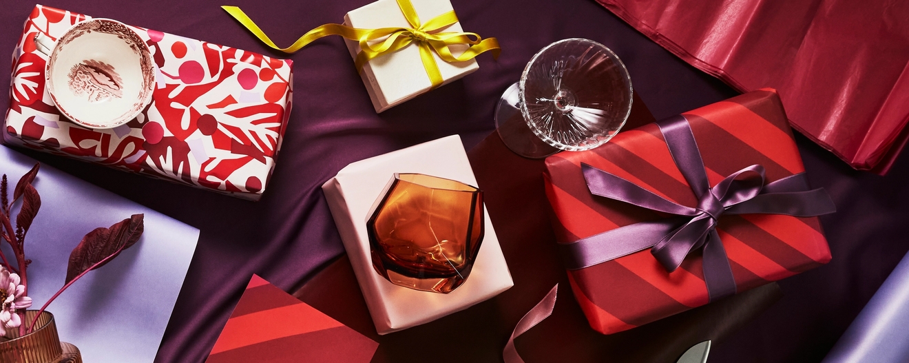 Gaveinnpakkede esker, glass og gavepapir i festlige røde, lilla og gulltoner.