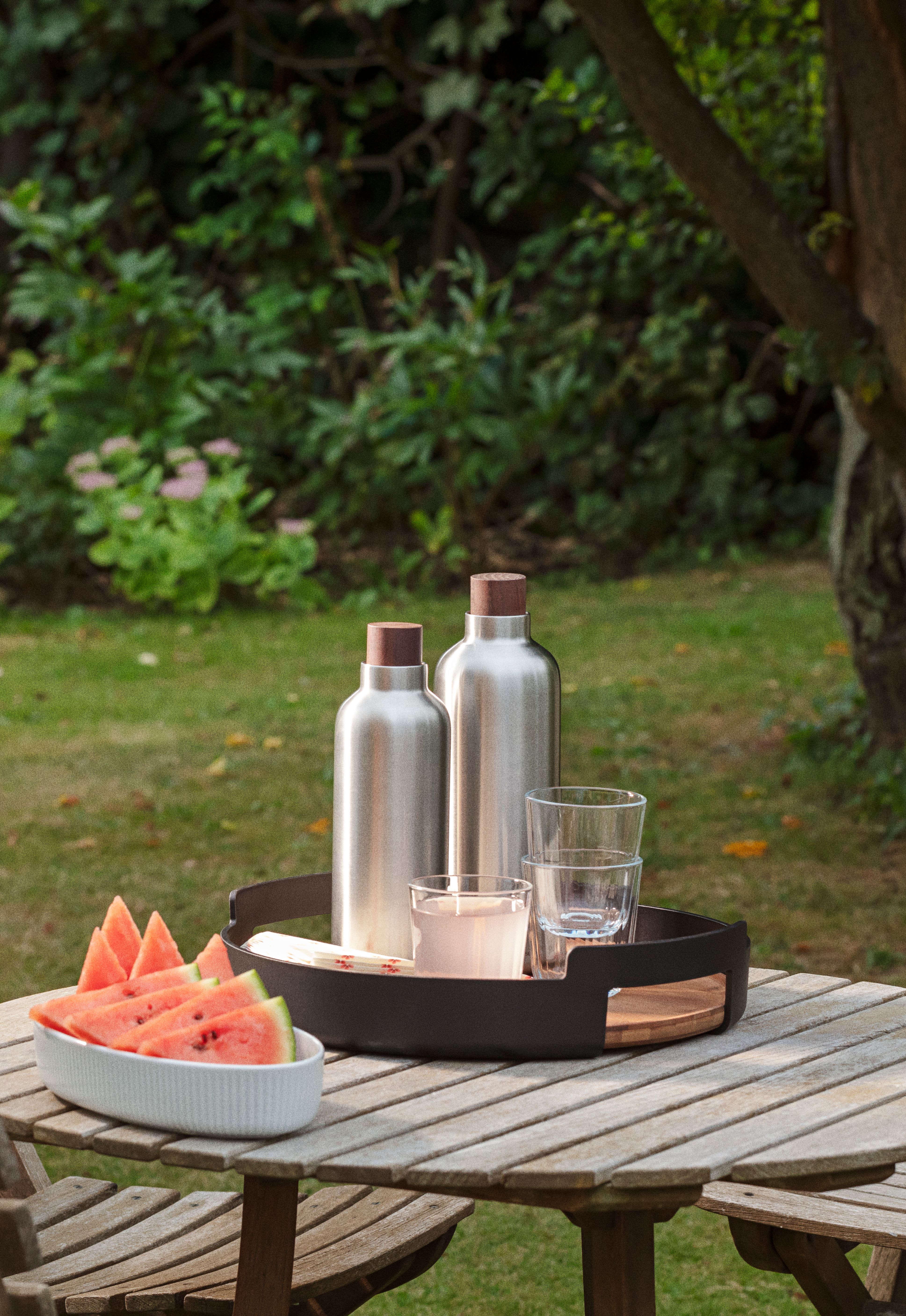 Vannmelon, to sølvfargede flasker og glass på et trebord i en hage