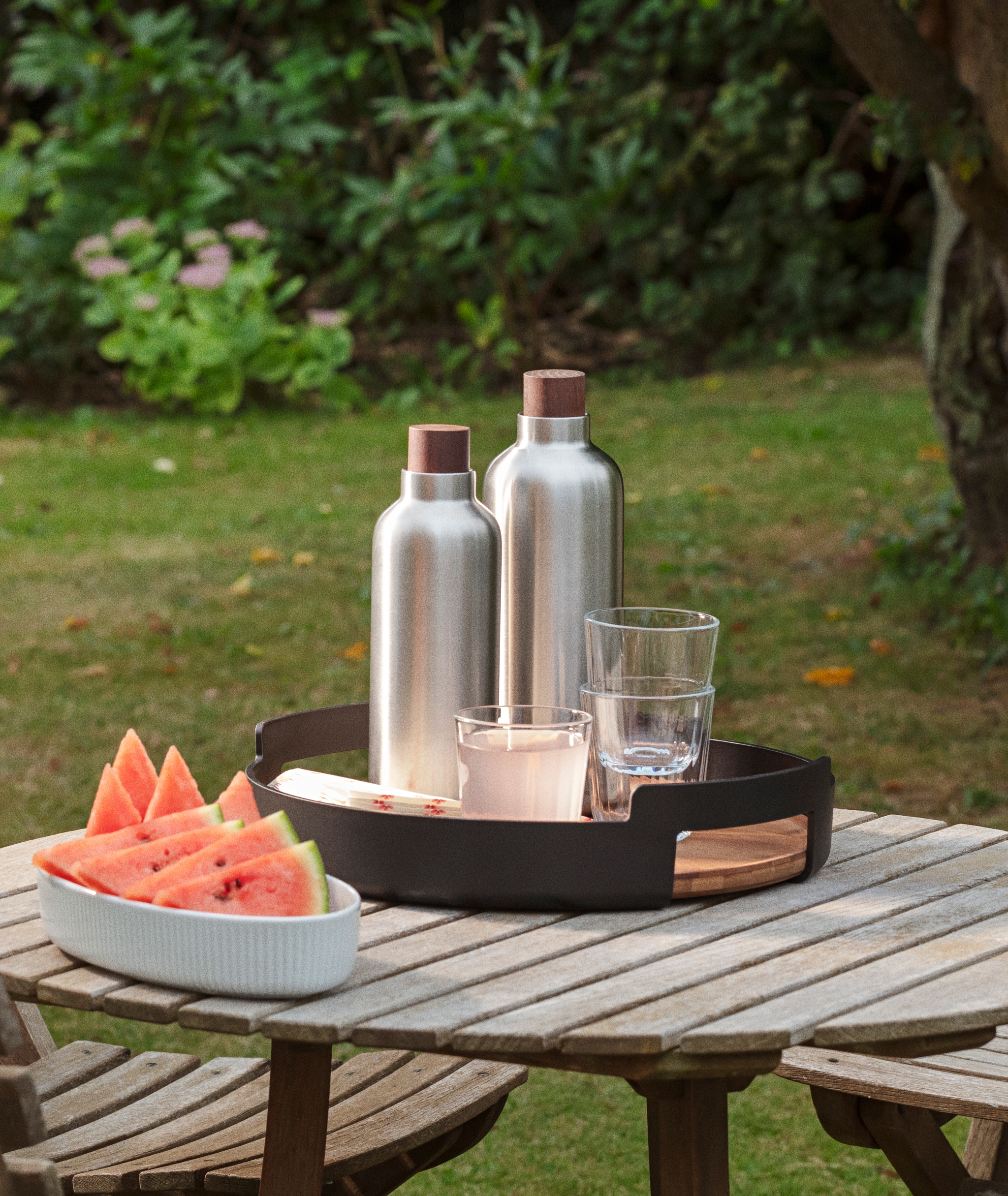Vattenmelon, två silverfärgade flaskor och glas på ett träbord i en trädgård