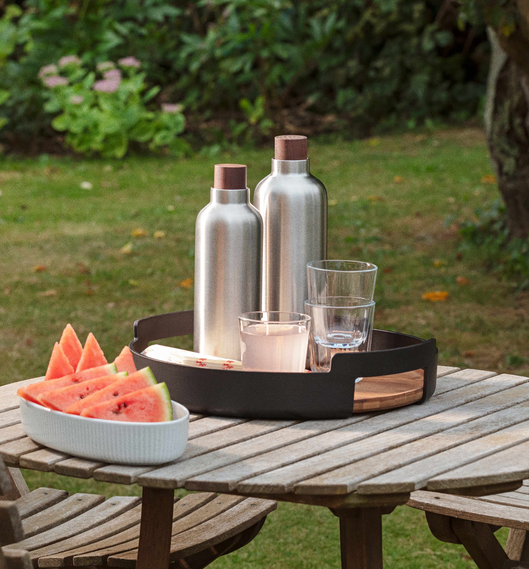 Vannmelon, to sølvfargede flasker og glass på et trebord i en hage