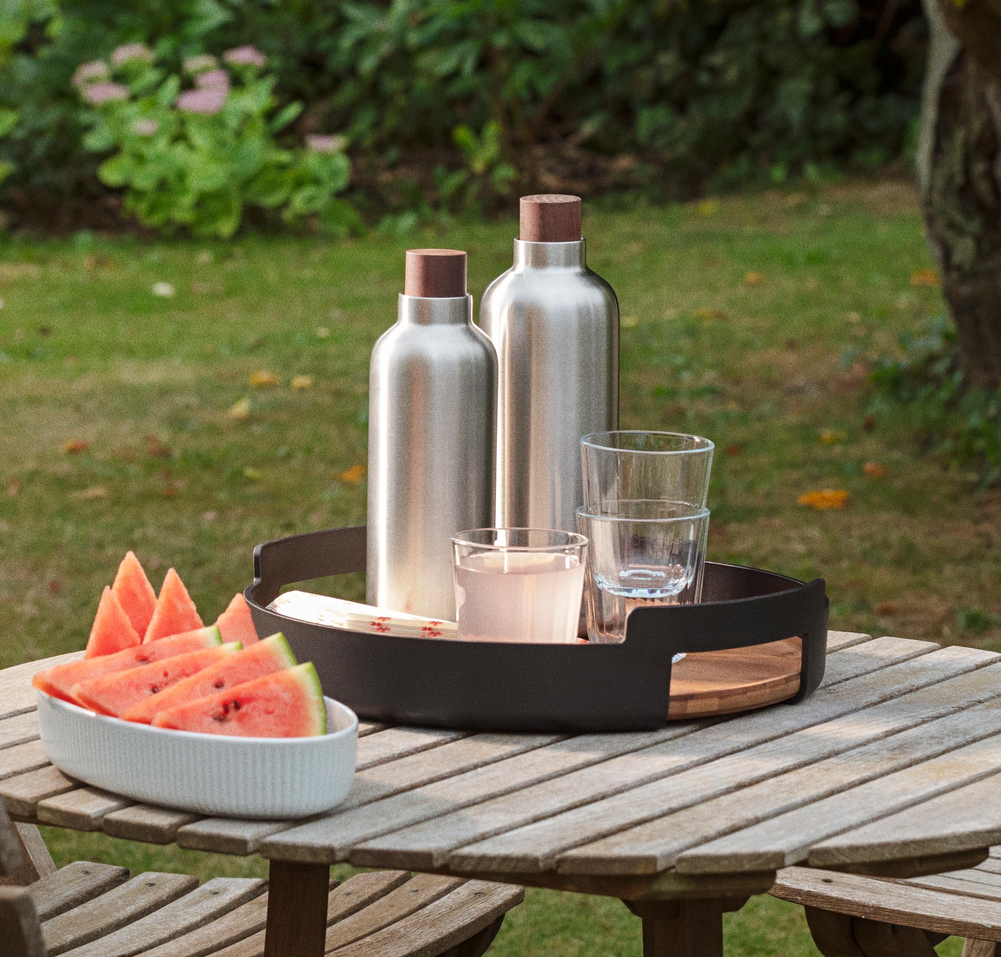 Vattenmelon, två silverfärgade flaskor och glas på ett träbord i en trädgård