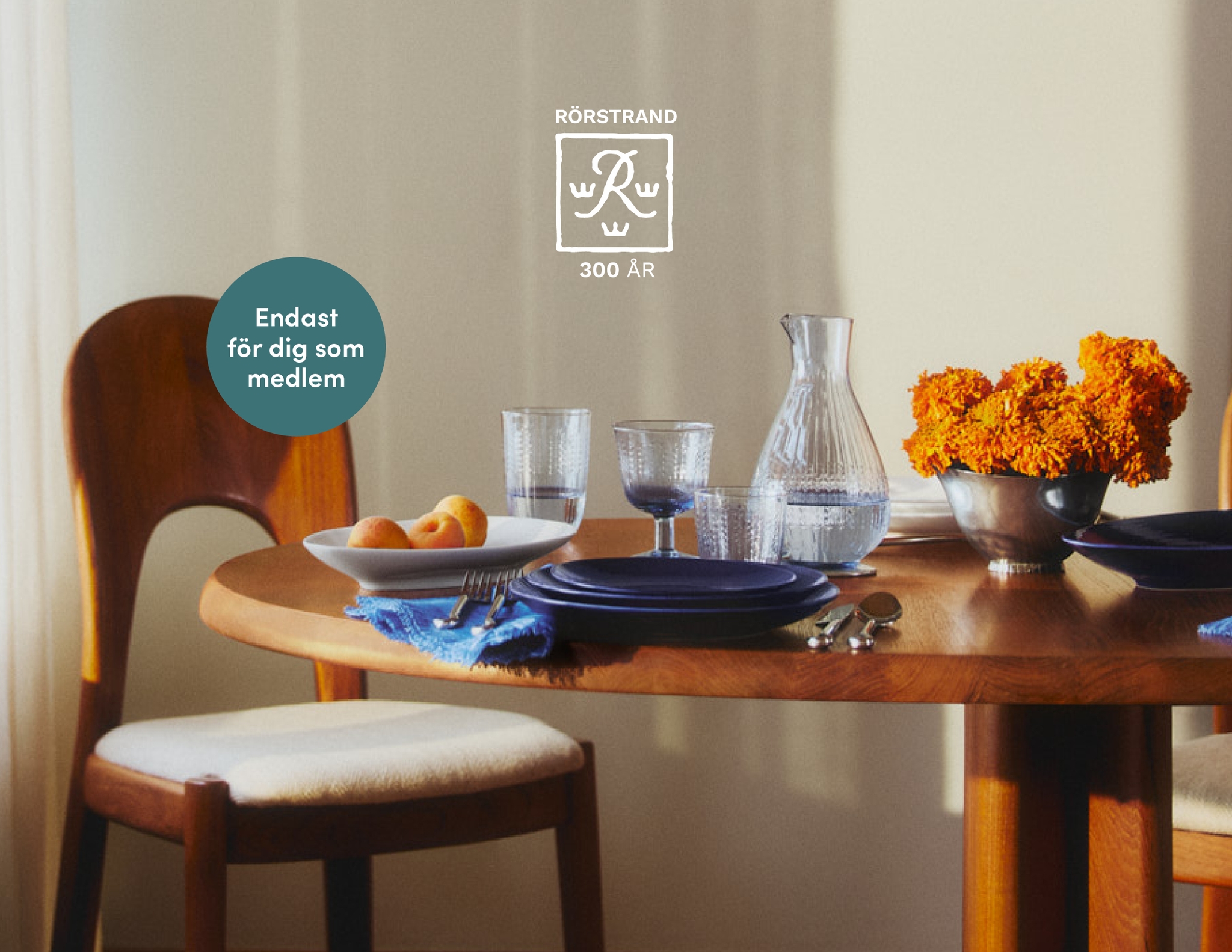 Ett matbord med mörkblå tallrikar, texturerade glas, en vattenkaraff, orange blommor och aprikoser. Text: RÖRSTRAND 300 ÅR och "Endast för dig som medlem".