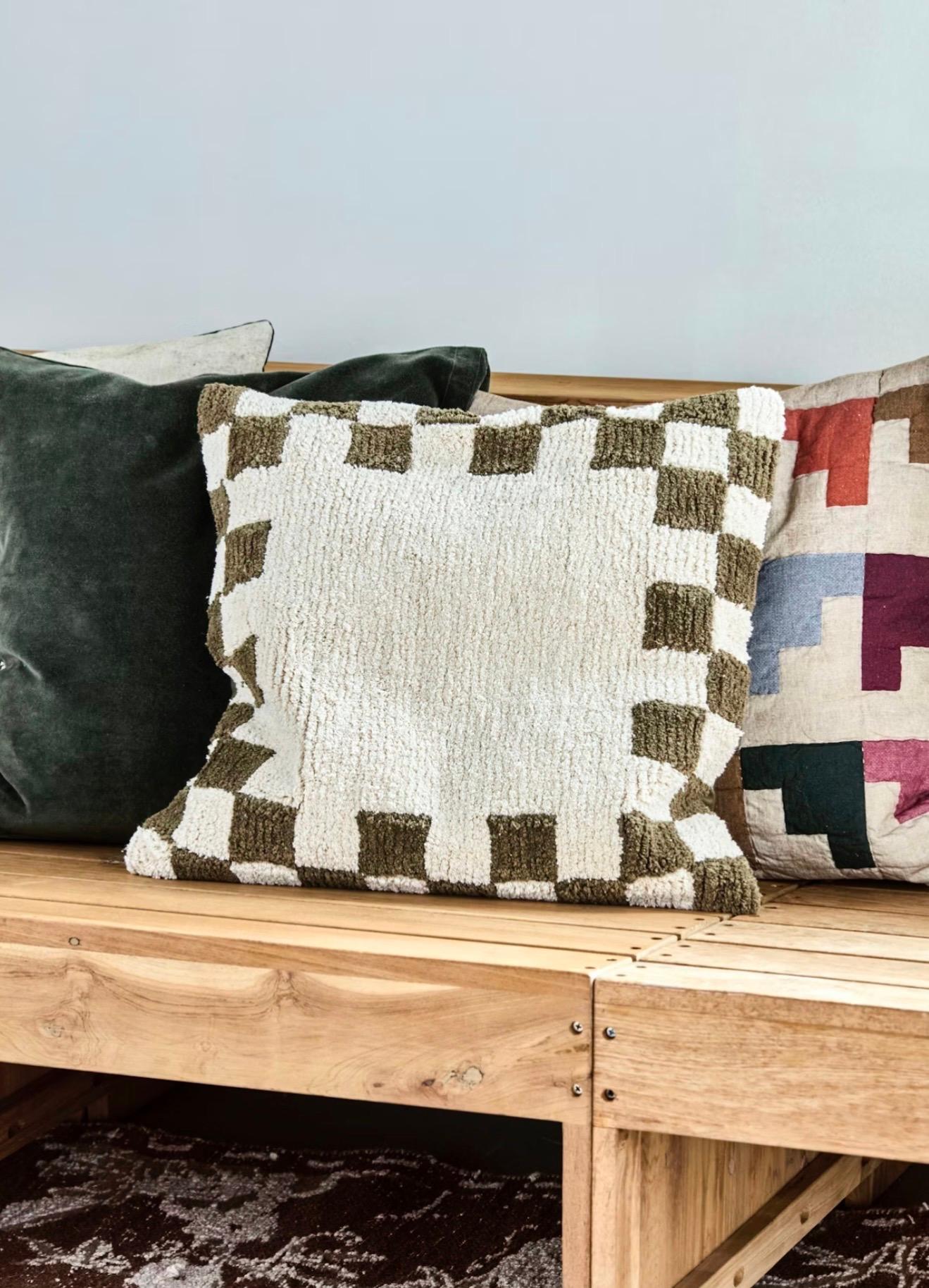 Three decorative pillows on a wooden bench: dark green velvet, cream with a brown checkerboard border, and multicolored patchwork.