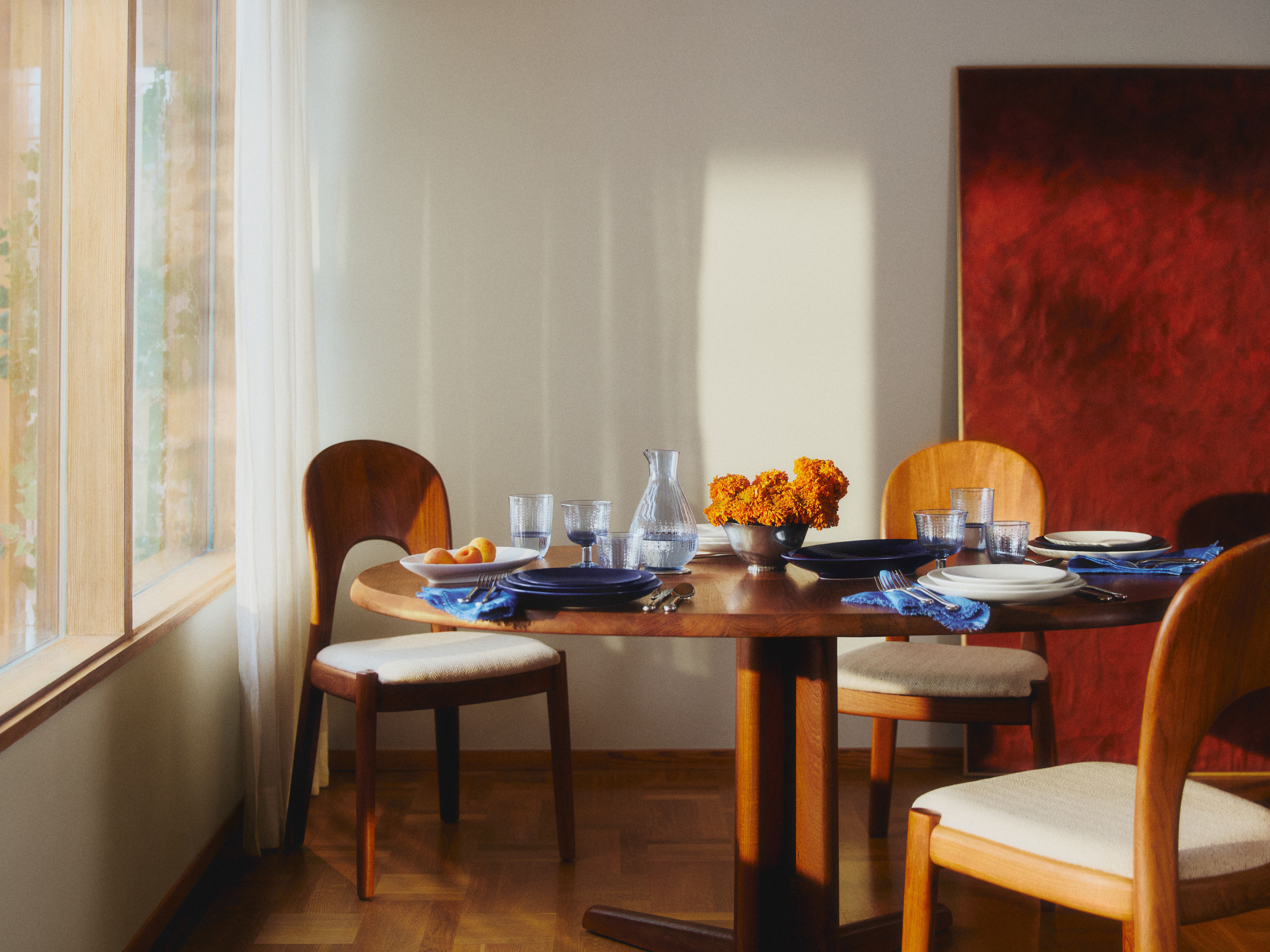 A round wooden dining table is set with blue plates, glasses, and orange flowers, surrounded by wooden chairs in a sunlit room with a large red abstract painting.