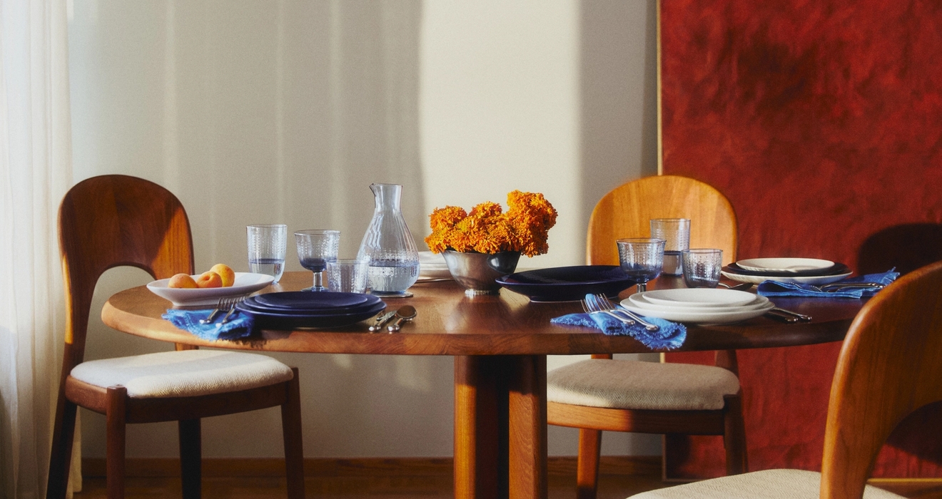 Ett runt matbord i trä är dukat med tallrikar, glas, blå servetter, orange blommor och en vattenkanna, upplyst av solljus från ett fönster, med ett rött abstrakt konstverk på väggen.