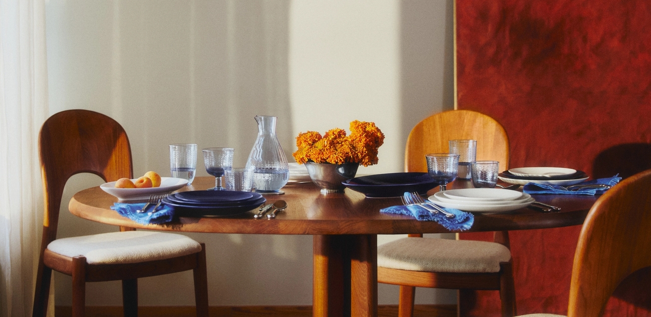 Ett runt matbord i trä är dukat med tallrikar, glas, blå servetter, orange blommor och en vattenkanna, upplyst av solljus från ett fönster, med ett rött abstrakt konstverk på väggen.