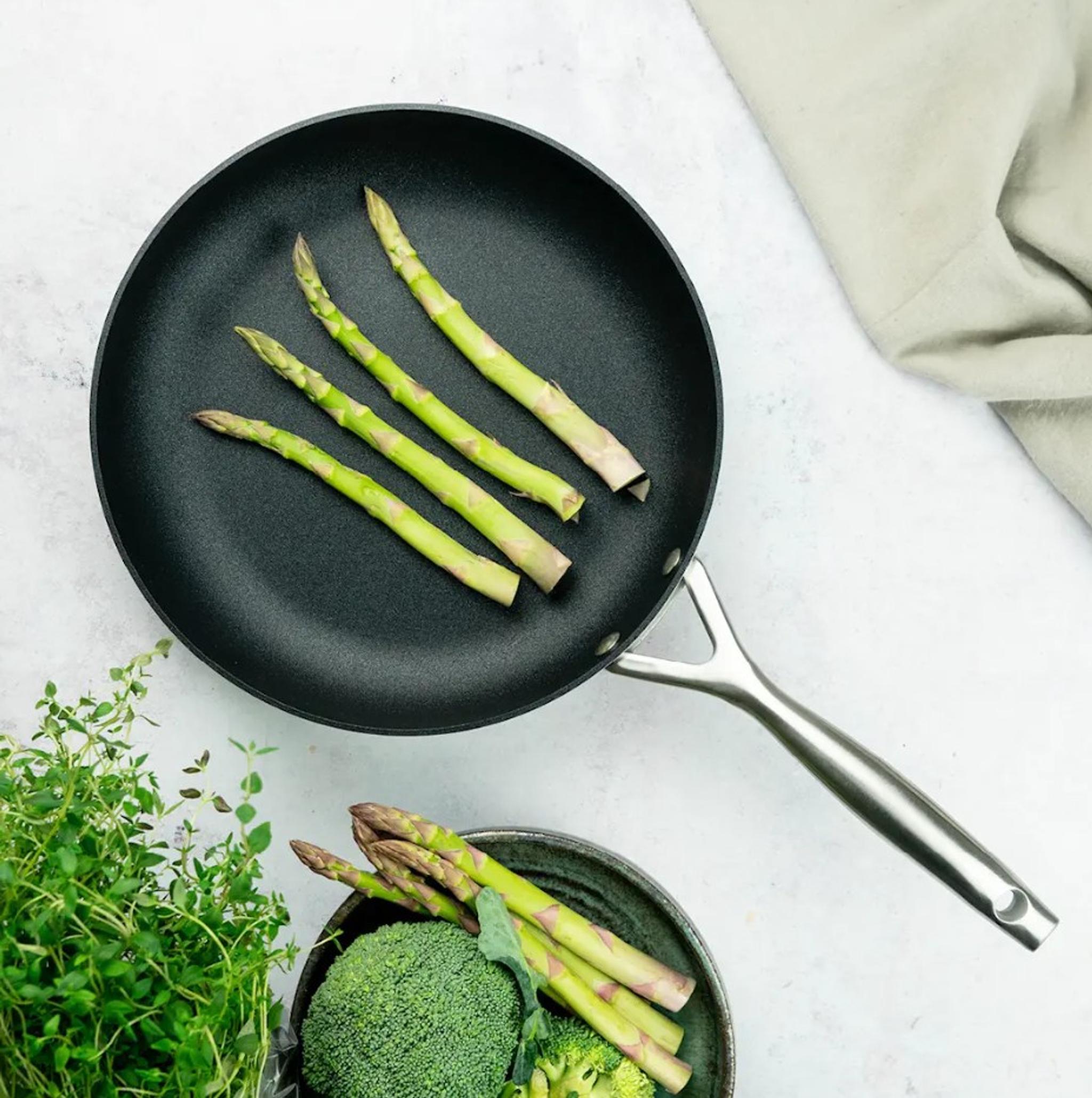 Grön sparris steks i en svart scanpan cs+ stekpanna bredvid färsk broccoli och örter.