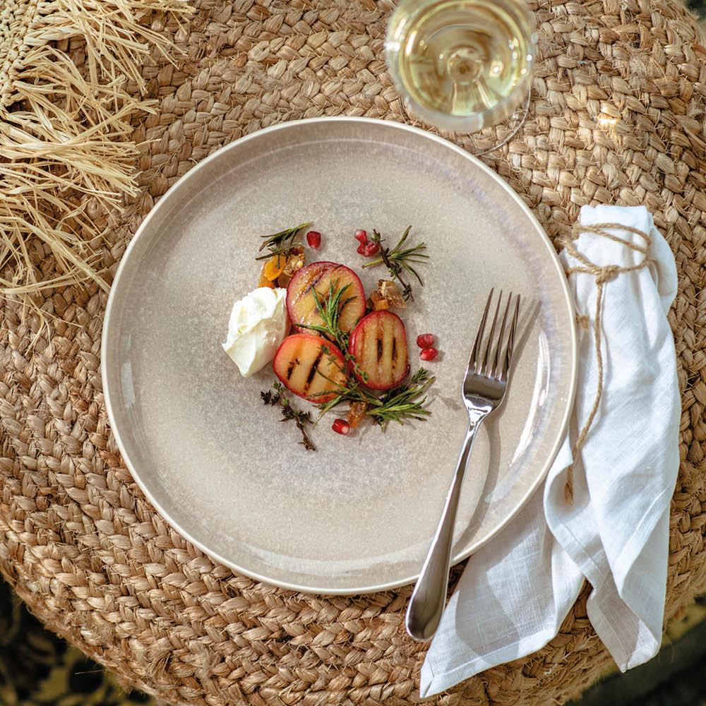 Tyylikkäästi aseteltu annos grillattuja hedelmiä, yrttejä ja kermavaahtoa vaalealla lautasella punotulla pöydällä viinilasin kera.