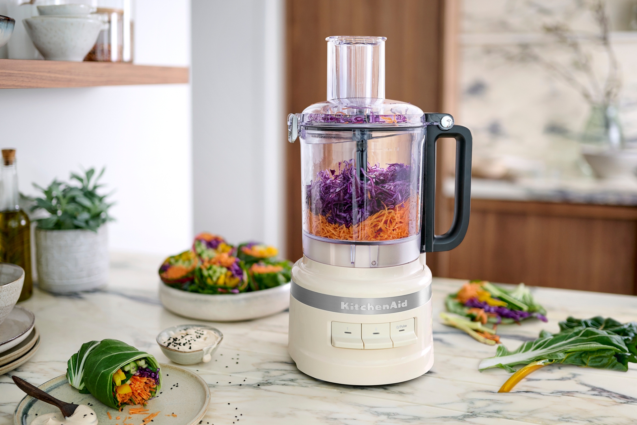 Matberedare från KitchenAid på köksbänk som river morötter och rödkål bredvid färgglada grönsakswraps.