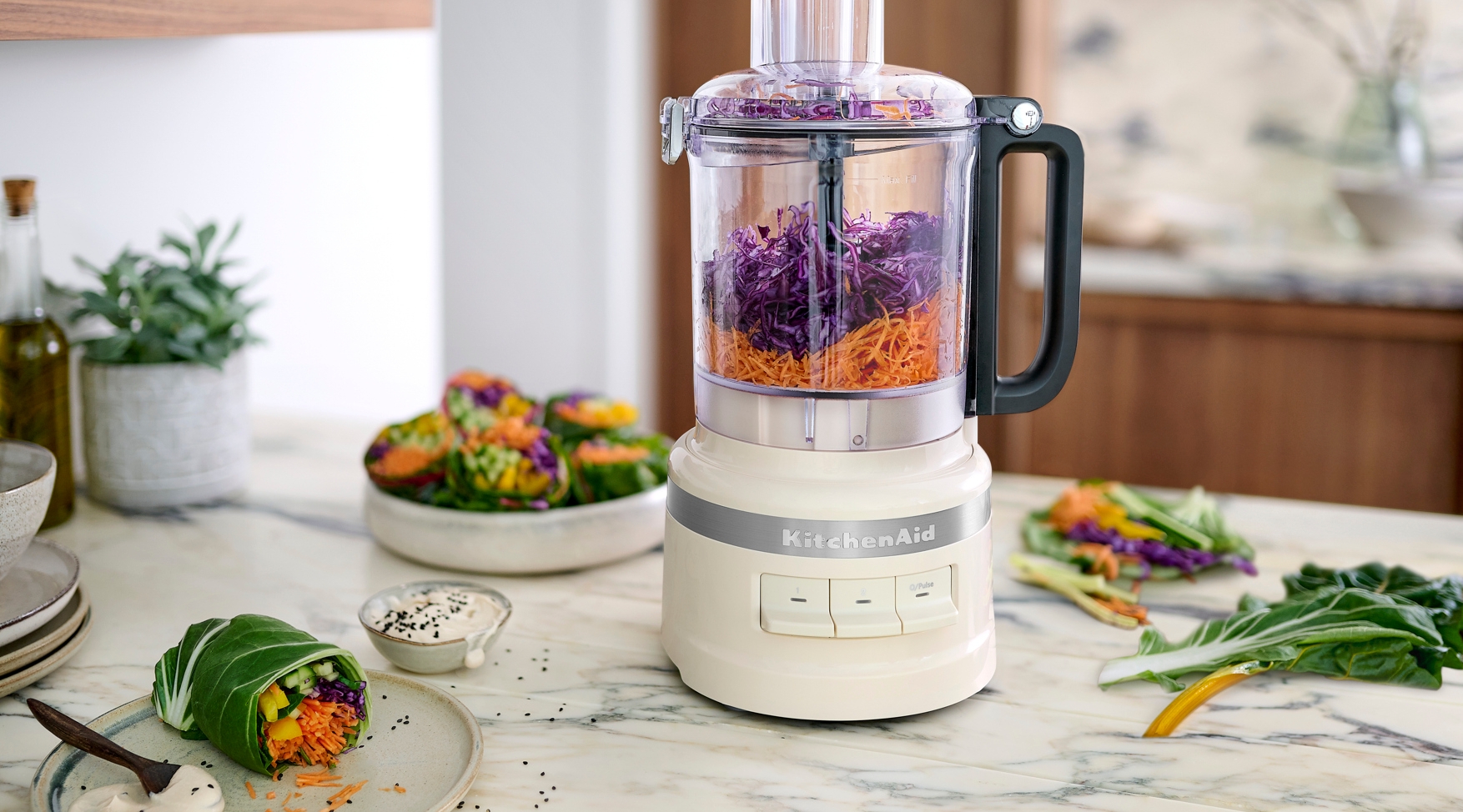 Kjøkkenmaskin fra KitchenAid hakker lilla kål og gulrøtter på en lys kjøkkenbenk med friske grønnsaker rundt.